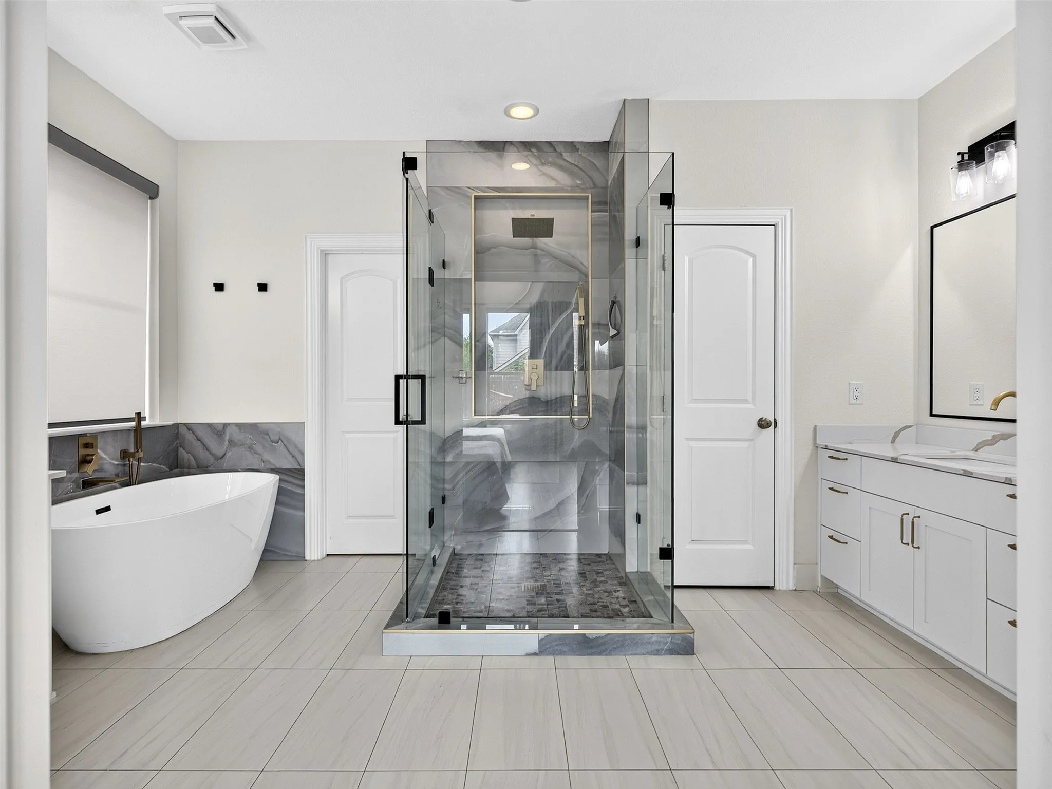 Spacious bathroom featuring a frameless glass shower enclosure with a patterned tile floor, a freestanding soaking tub with wall-mounted bronze-tone fixtures, a dual-sink vanity with white cabinetry and gold-tone faucets, and light-toned tile flooring