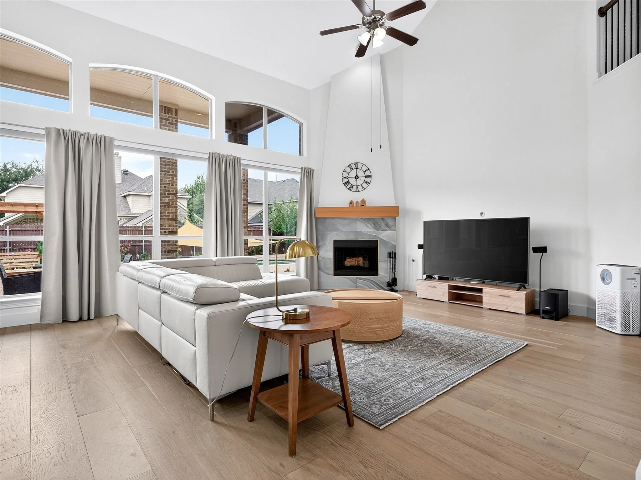 Soaring ceilings with a ceiling fan, a fireplace with a wood mantle and tiled surround, wood-finish flooring, and expansive windows with arched transoms