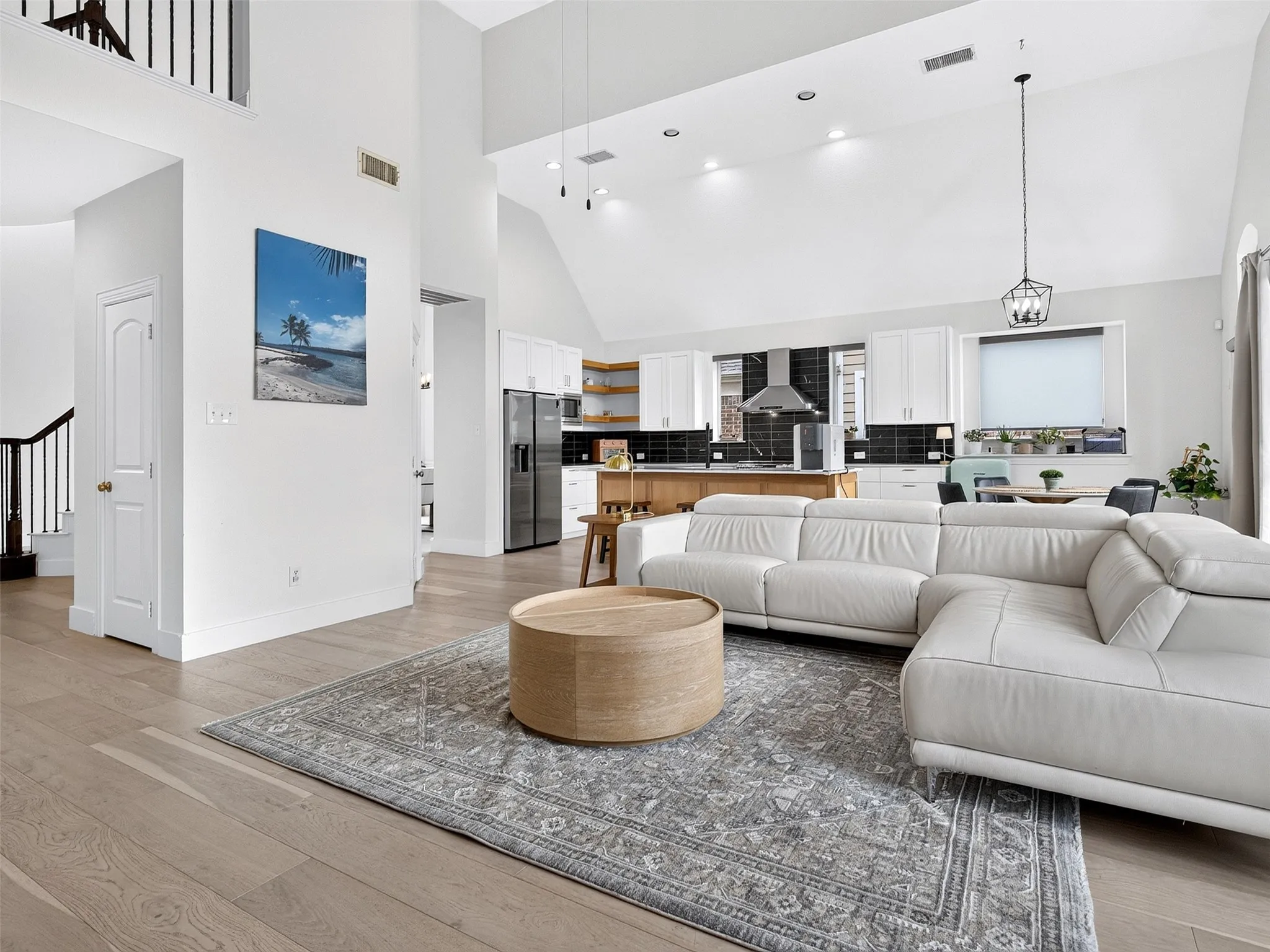 Spacious open-concept living area featuring light wood-finish flooring, a vaulted ceiling, recessed lighting, and a contemporary kitchen with white cabinetry and dark tile backsplash