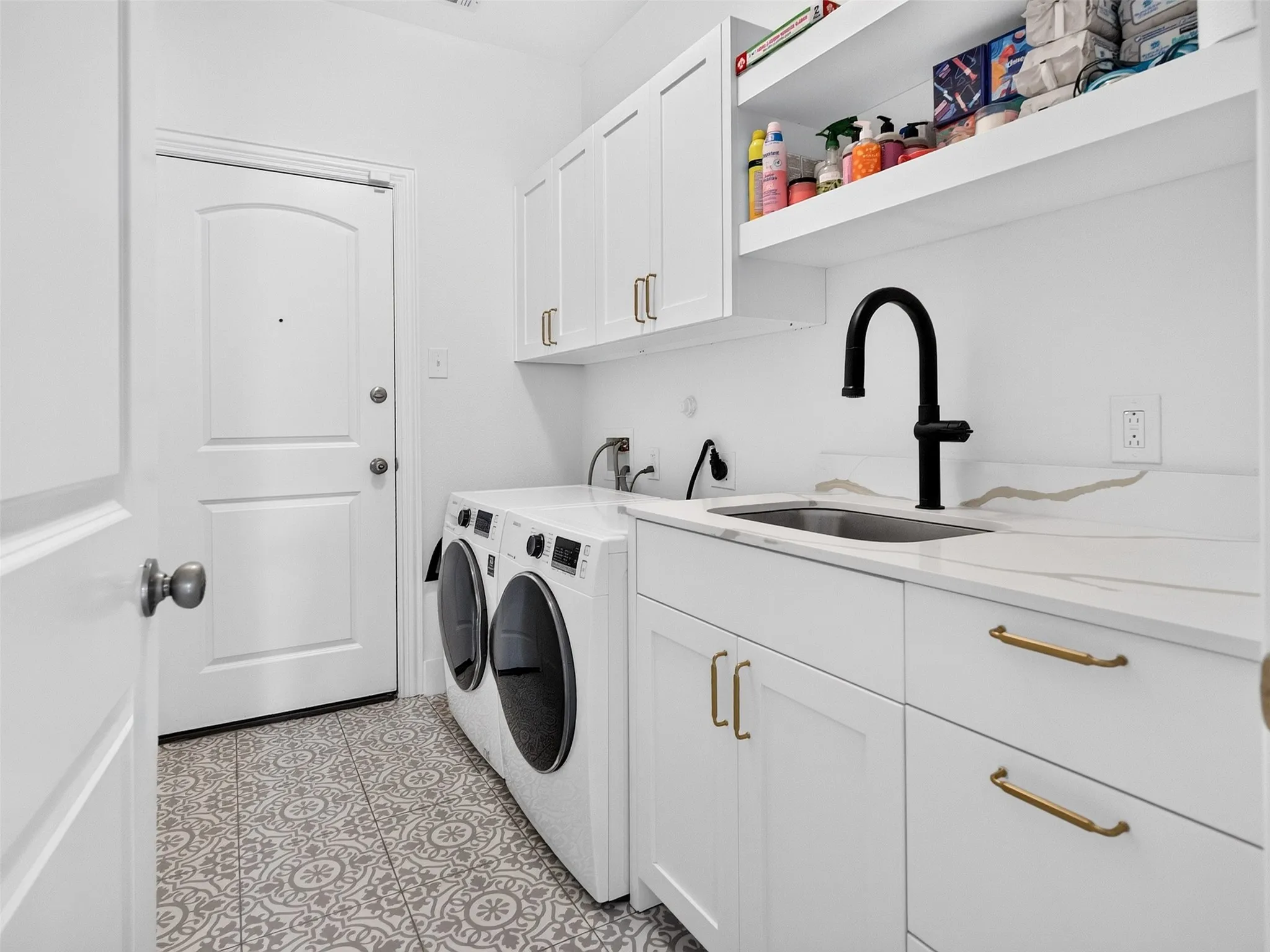 Laundry room featuring patterned floor tiles, white shaker cabinetry with brass hardware, integrated sink with matte black faucet, and open shelving