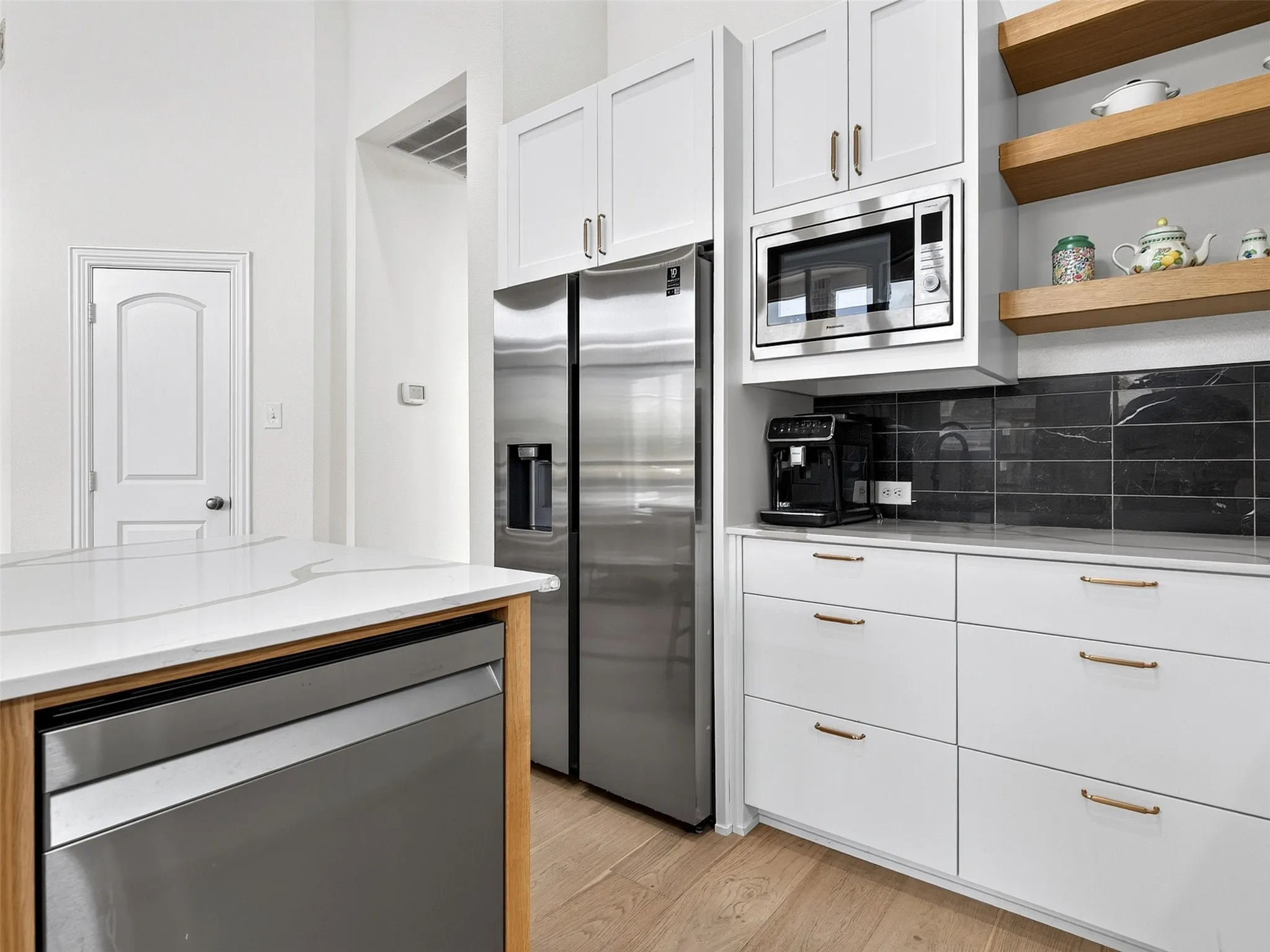 Modern kitchen featuring a stainless steel refrigerator, built-in microwave, white cabinetry with gold-tone hardware, a dark subway tile backsplash, and light wood-finish flooring