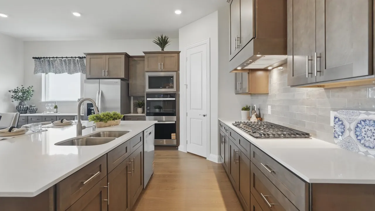 Gourmet kitchen featuring a large island with dual stainless steel sinks, built-in stainless steel appliances, wood-finish cabinetry, light-toned countertops, and a subway tile backsplash