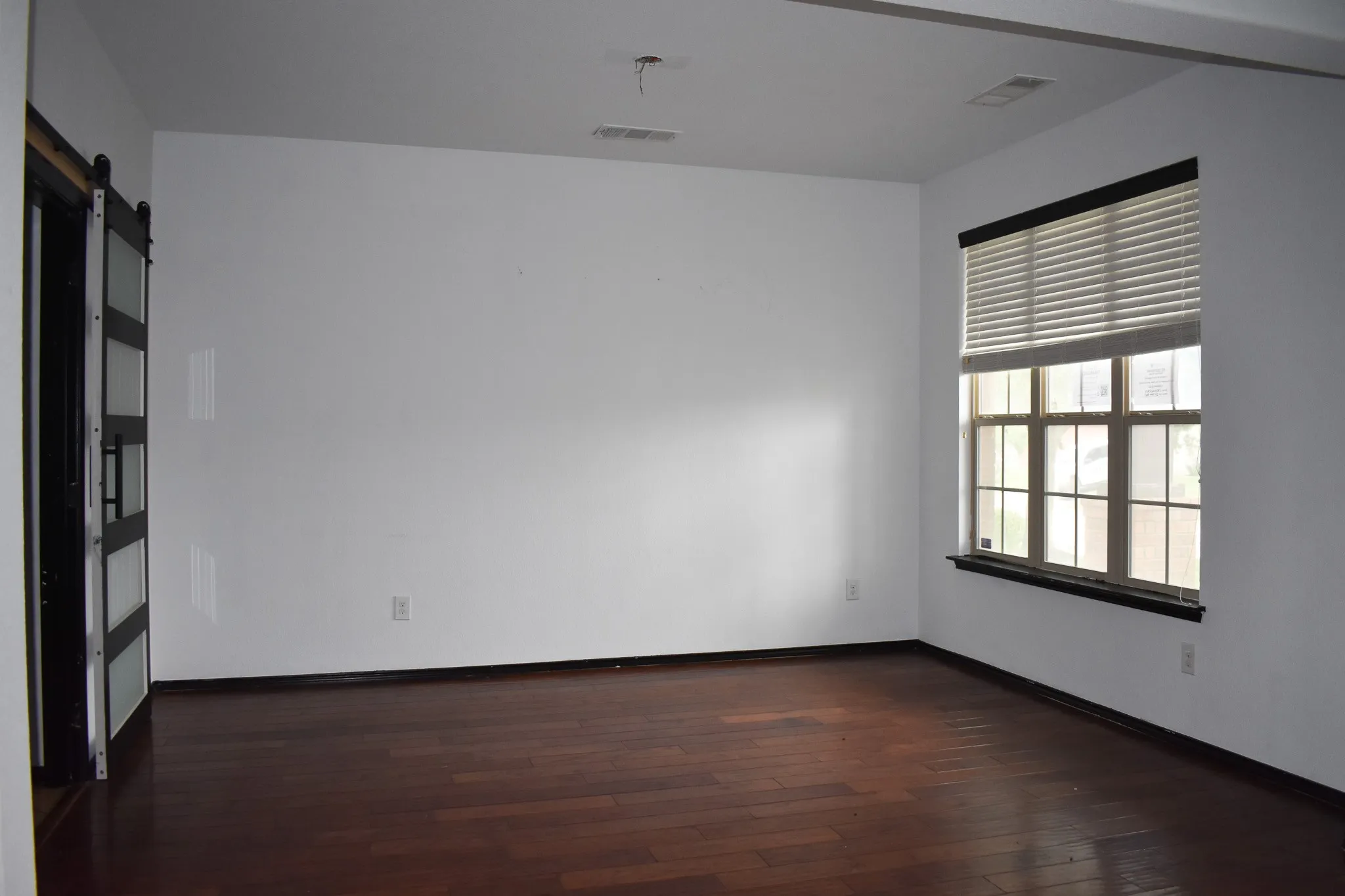 Spacious interior room featuring wood-finish flooring, a large multi-pane window with blinds, white walls, and a modern barn-style door with frosted glass panels