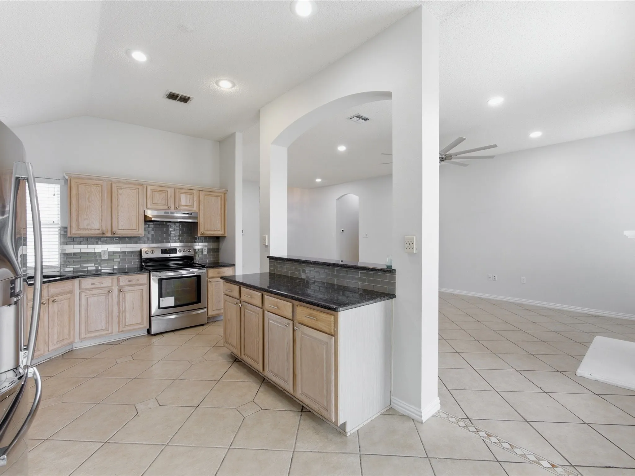 Kitchen featuring light wood cabinetry, dark countertops, stainless steel appliances, a tile backsplash, and tile flooring