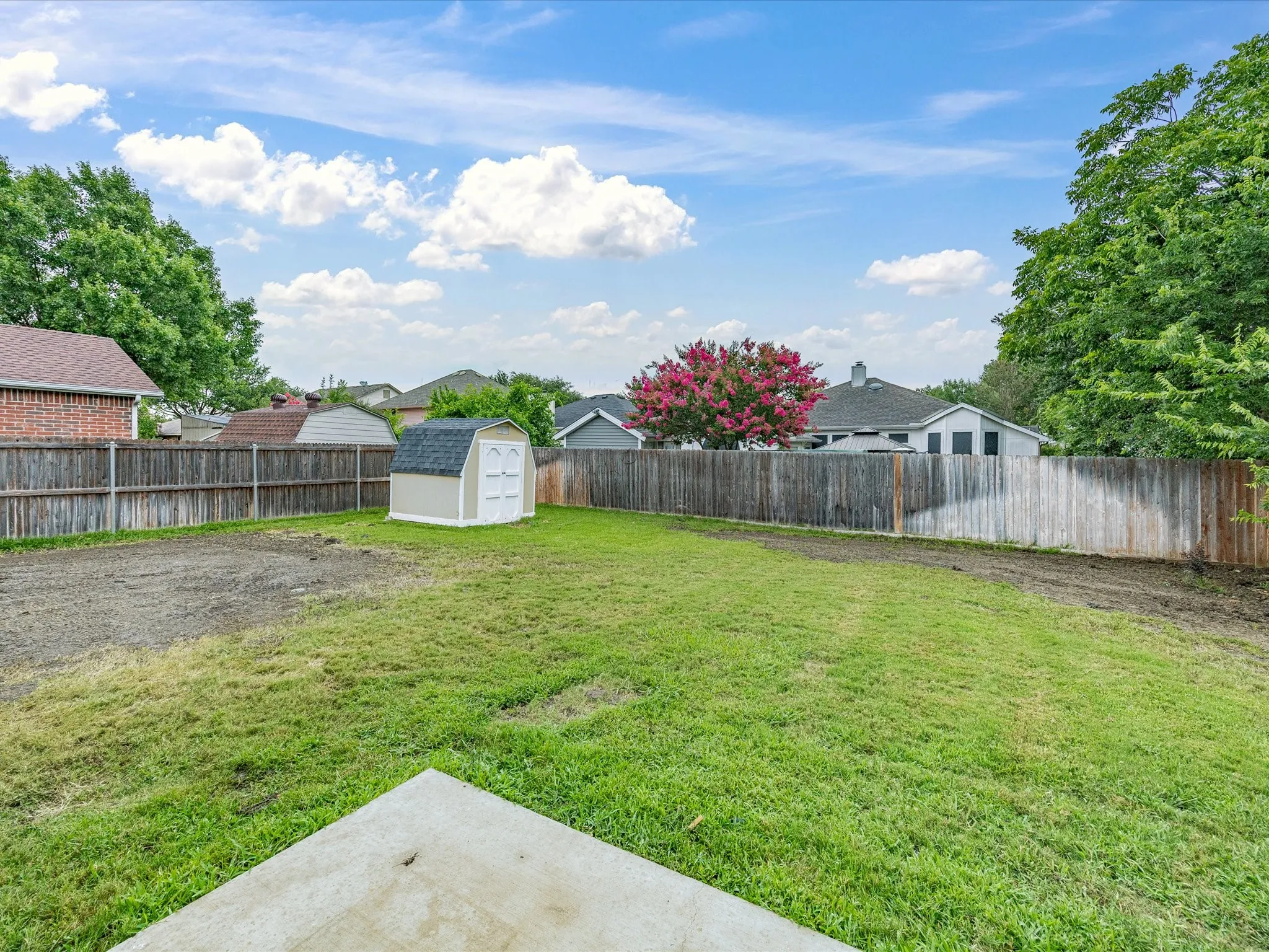 Expansive backyard featuring a combination of grass and bare earth, wood privacy fencing, a storage shed, and a concrete patio section