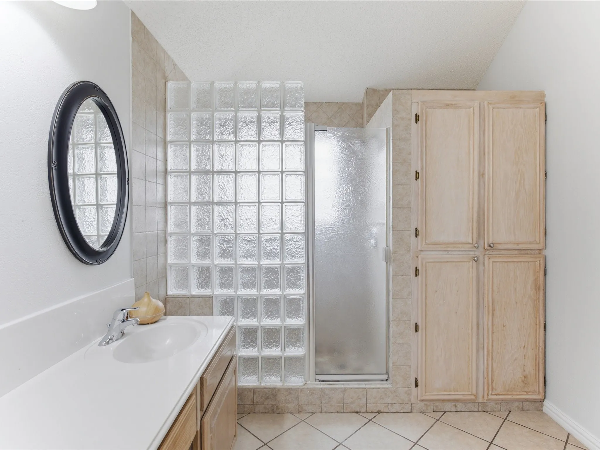 Bathroom vanity with an oval mirror, glass block shower enclosure, frosted glass shower door, and built-in storage cabinetry