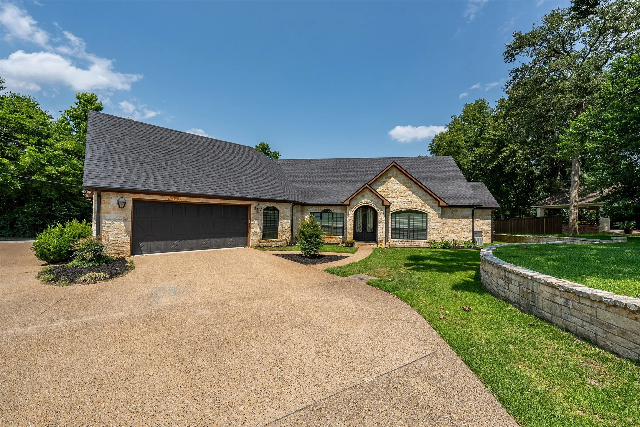 Stone facade residence featuring an expansive driveway, a two-car garage, and a dark shingle roof