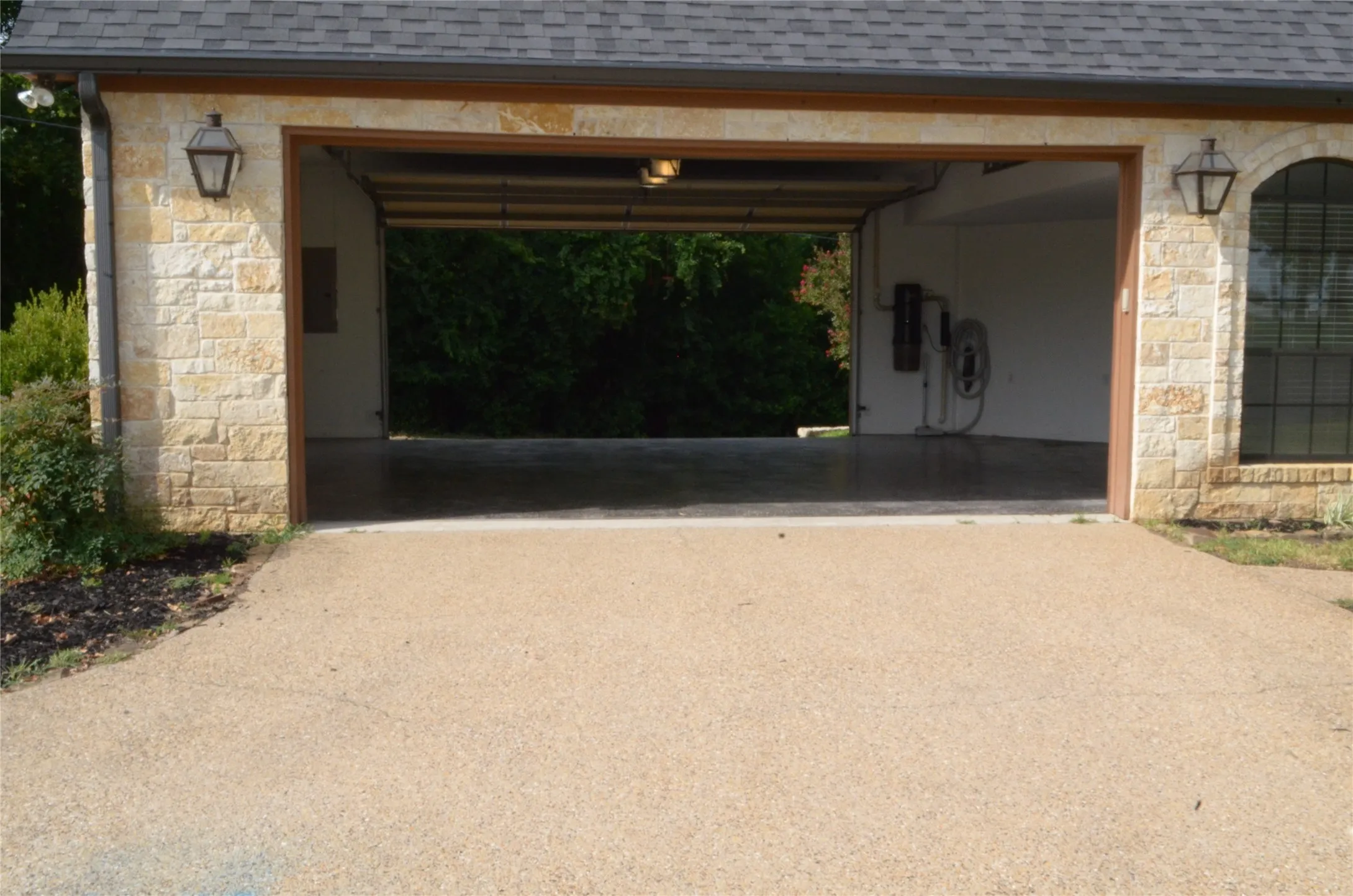 Exposed aggregate driveway leading to a two-car garage with stone veneer exterior, architectural shingle roof, and carriage-style outdoor lighting fixtures