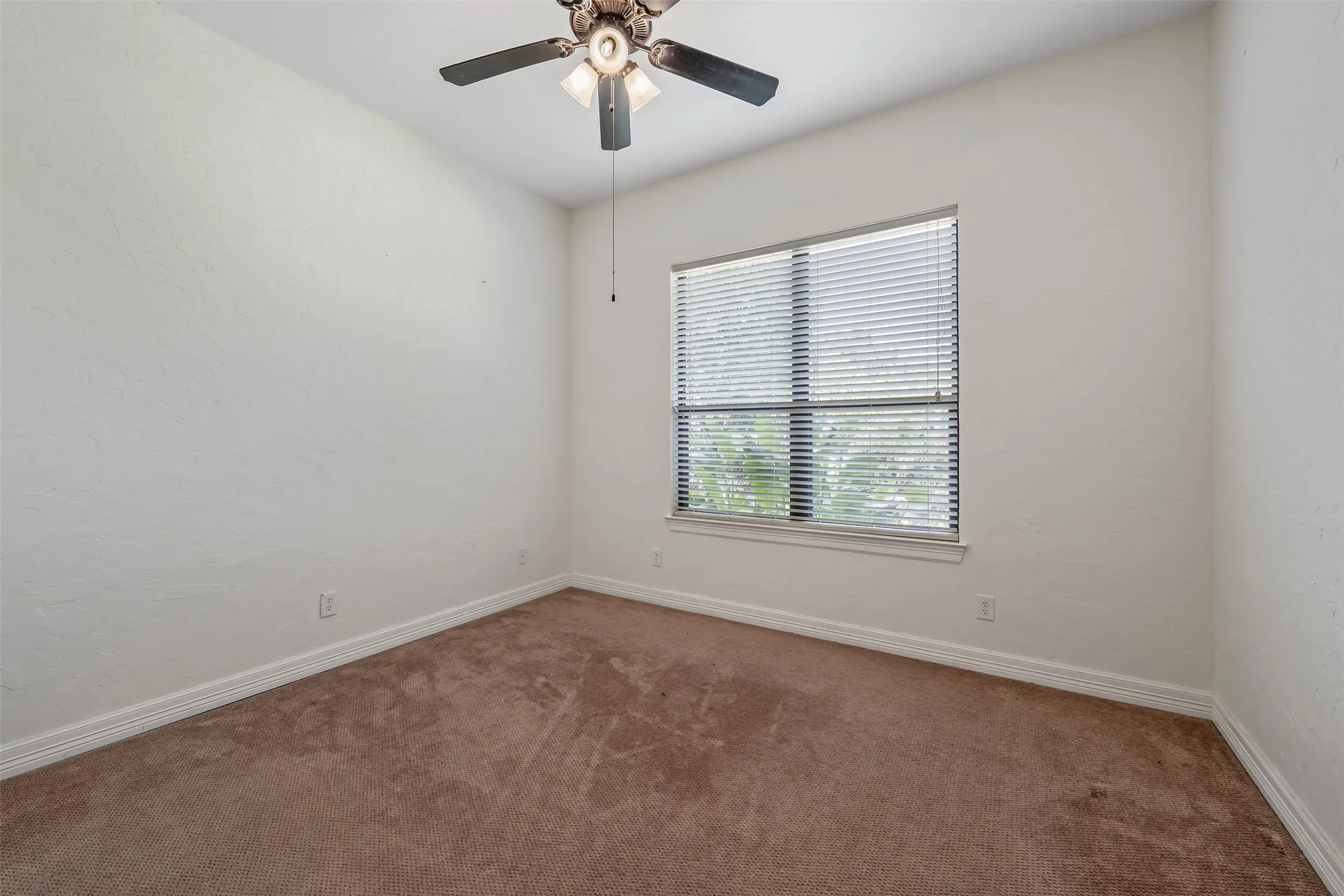 Carpeted room featuring a ceiling fan with integrated lighting, a window with horizontal blinds, textured walls, and white baseboards