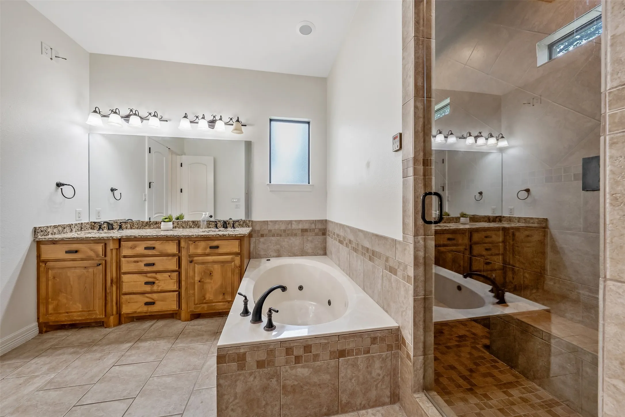 Bathroom featuring a jetted soaking tub, a tiled walk-in shower with a glass enclosure, dual vanity with wood cabinetry and stone countertops, and tile flooring