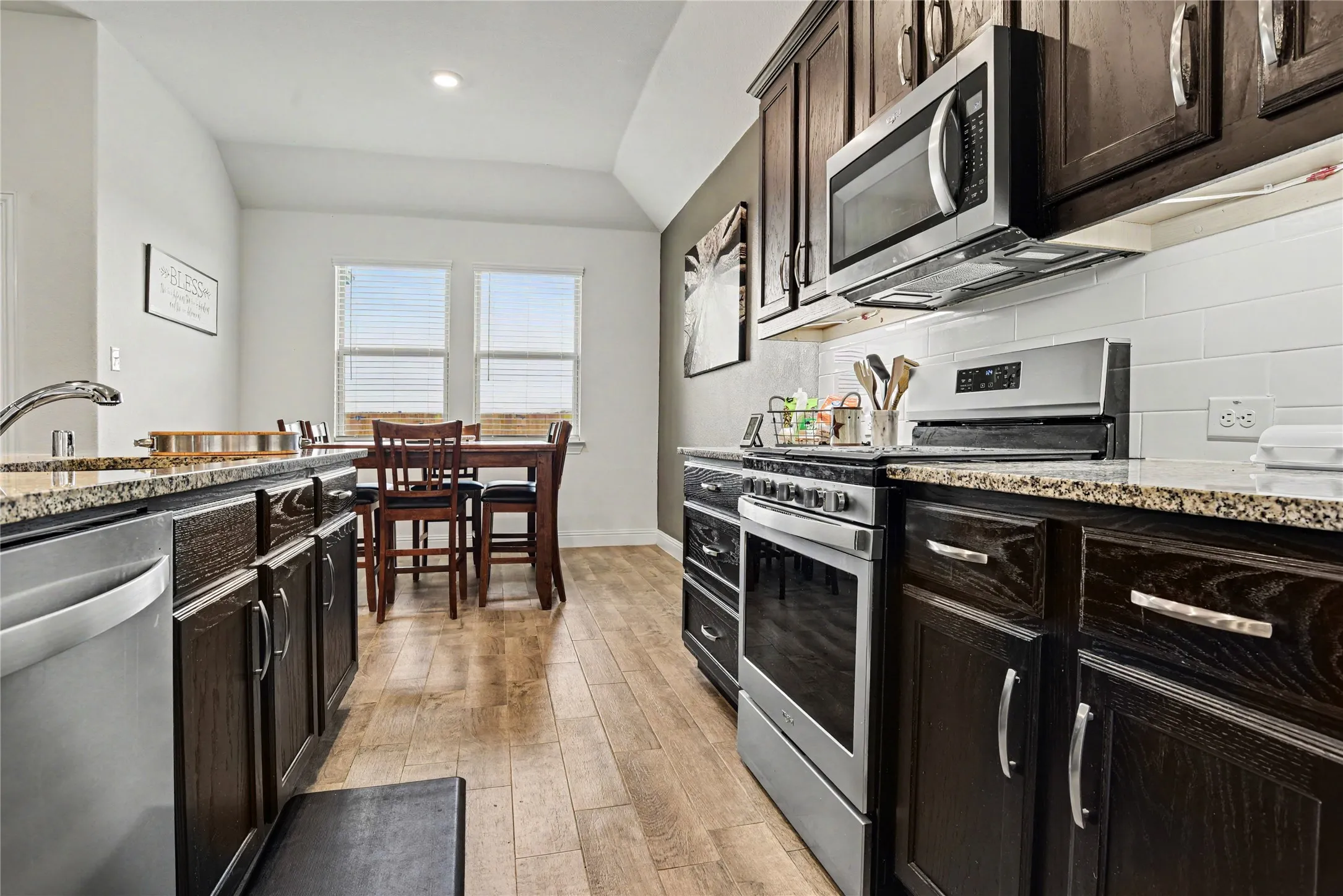 Kitchen featuring wood-finish flooring, dark wood cabinetry, granite-style countertops, stainless steel appliances, and a white subway tile backsplash