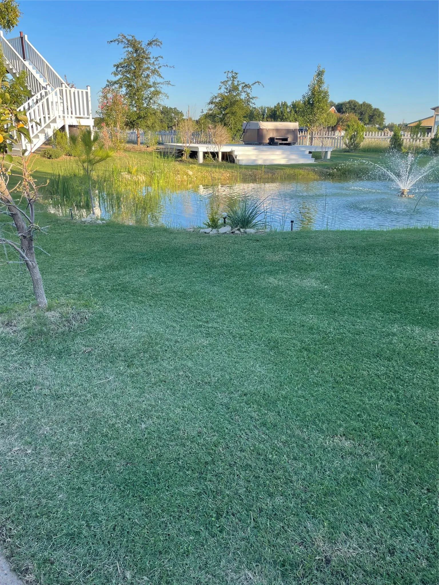 Expansive green lawn with a large pond, white deck, hot tub, and water fountain feature
