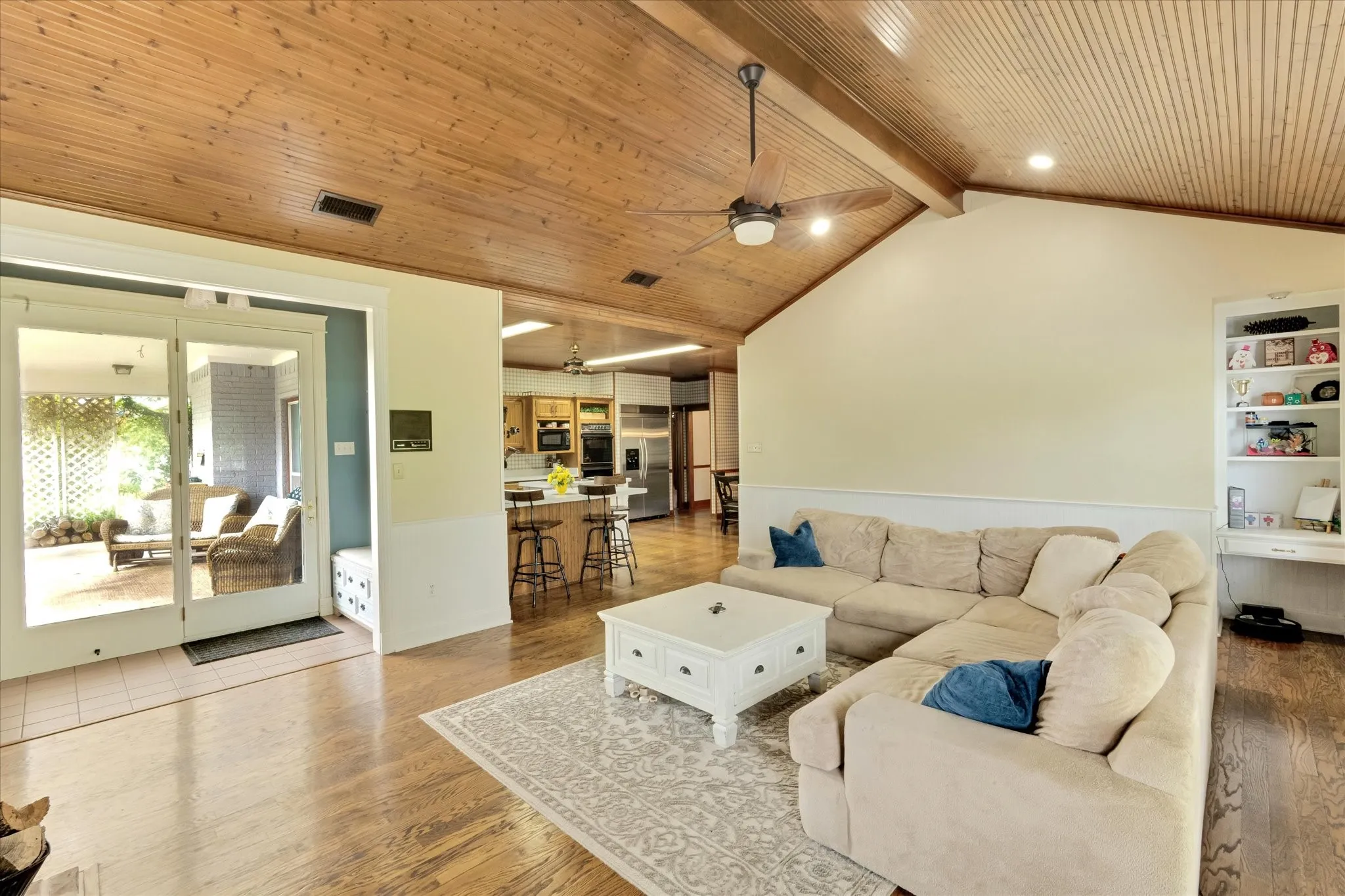 Living area featuring vaulted wood-paneled ceilings with exposed beams, wood-finish flooring, built-in shelving, and direct access to an enclosed patio through glass doors
