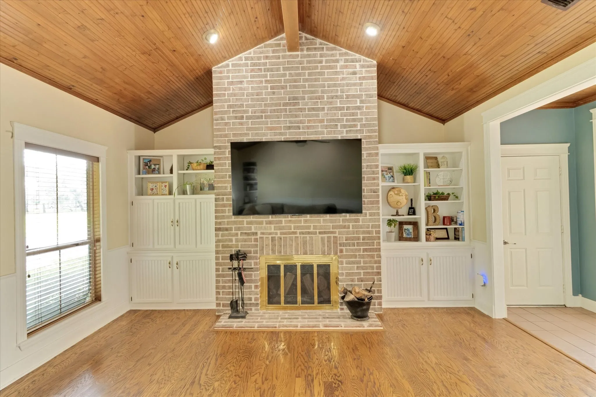 Vaulted wood-paneled ceiling with exposed beam, a brick fireplace surround, built-in shelving, wood-finish flooring, and a large window with blinds