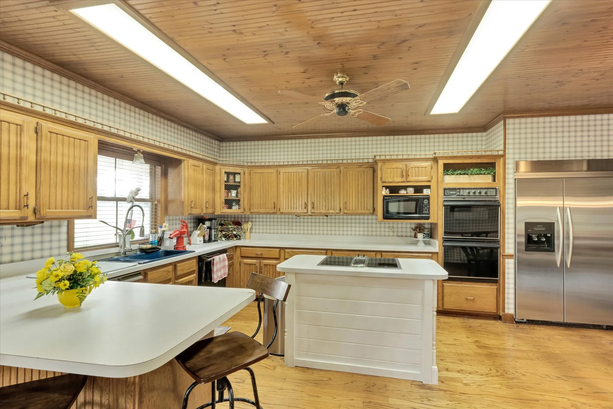 Kitchen featuring wood-finish flooring, wood-paneled ceiling, light wood cabinetry, stainless steel appliances, and a central island with an integrated cooktop. Sub-Zero built in refrigerator