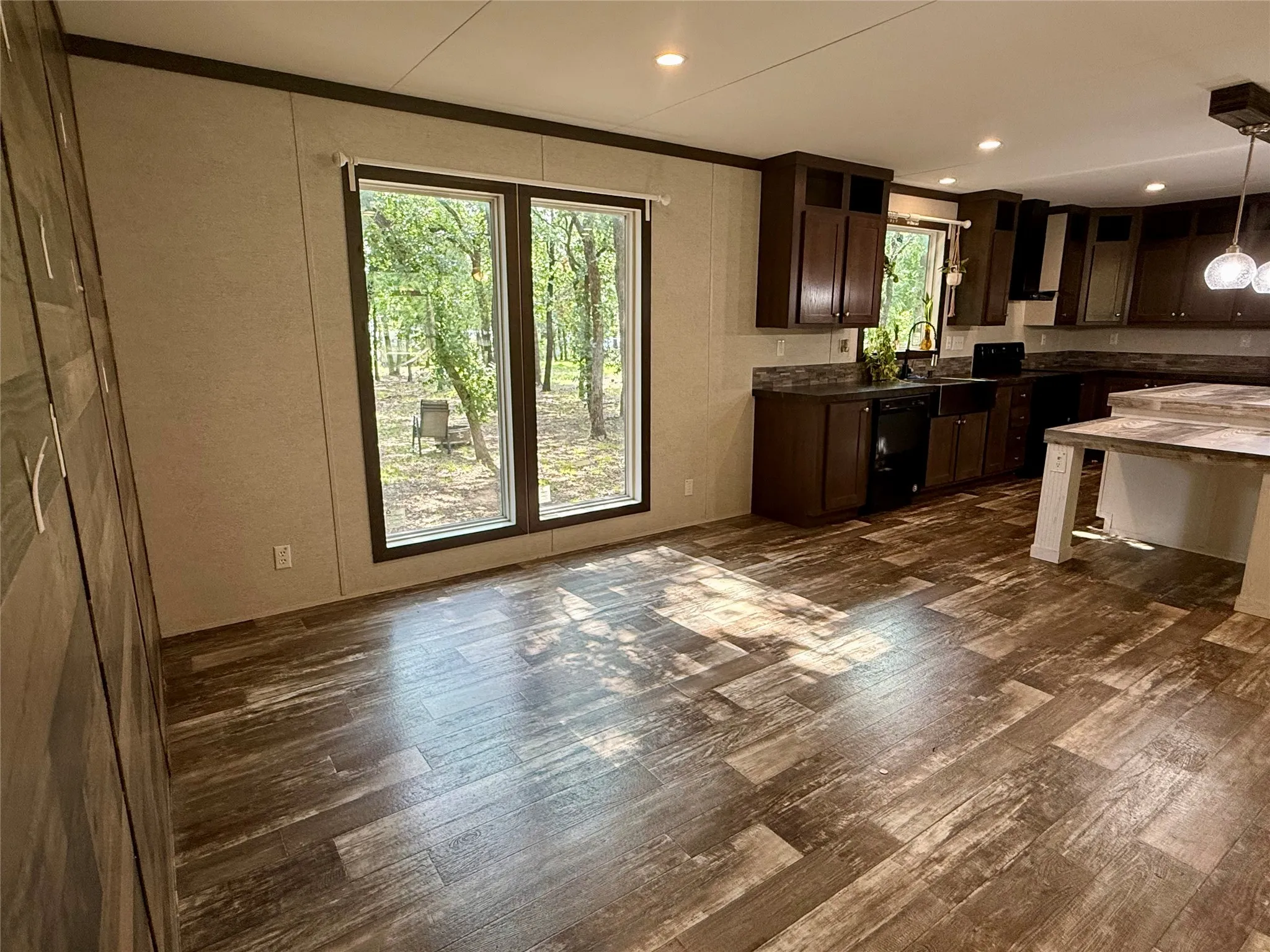 Open-concept kitchen and living area featuring wood-finish flooring, dark wood cabinetry, a built-in island with countertop, recessed lighting, and large windows overlooking exterior greenery