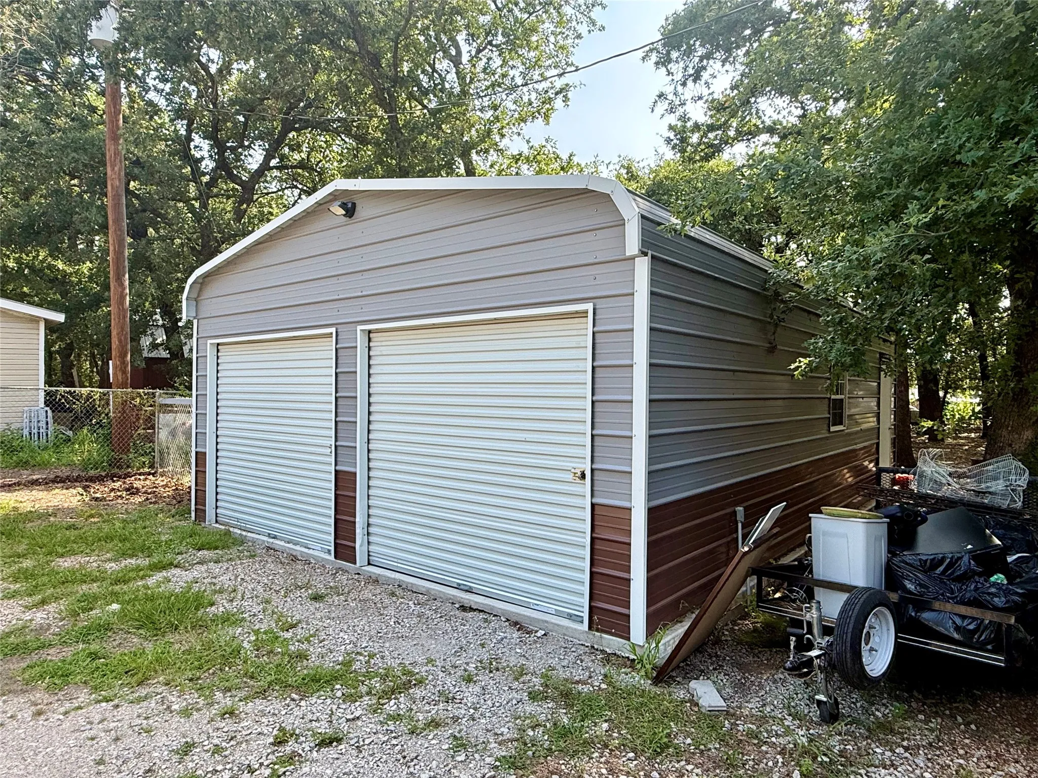 Two-bay metal structure with roll-up doors, a side window, and exterior lighting, set on a gravel and grass surface