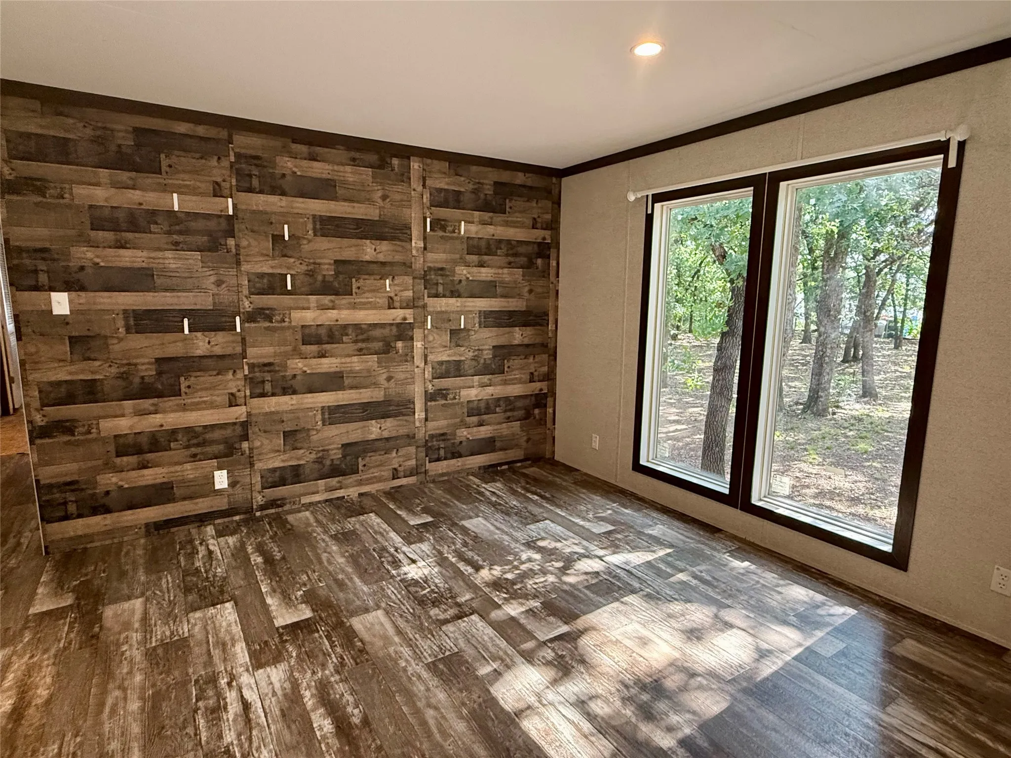 Spacious room featuring wood-finish flooring, a prominent accent wall with varied wood paneling, and two large windows with dark trim providing ample natural light
