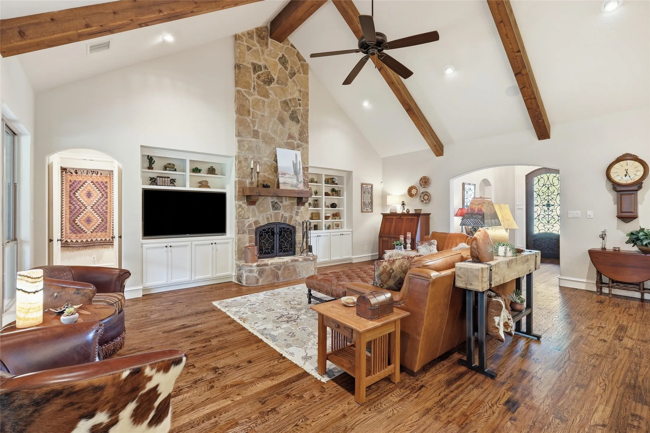 Living area featuring a floor-to-ceiling stone fireplace with a wood mantel, vaulted ceiling with exposed wood beams, wood-finish flooring, and built-in shelving with lower cabinetry
