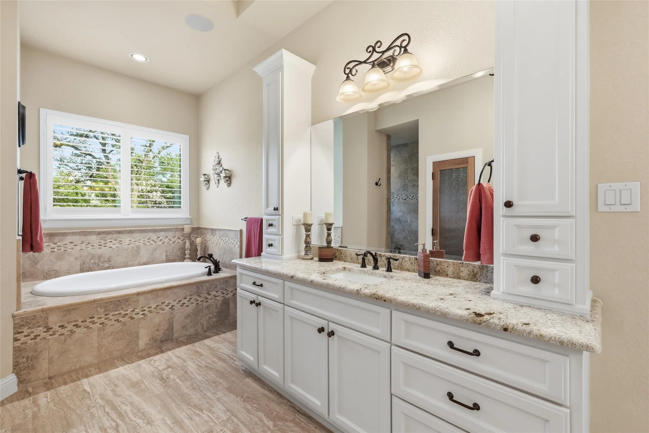 Spacious bathroom featuring an oval soaking tub with tile surround, plantation shutters, a dual vanity with granite countertop, and white cabinetry