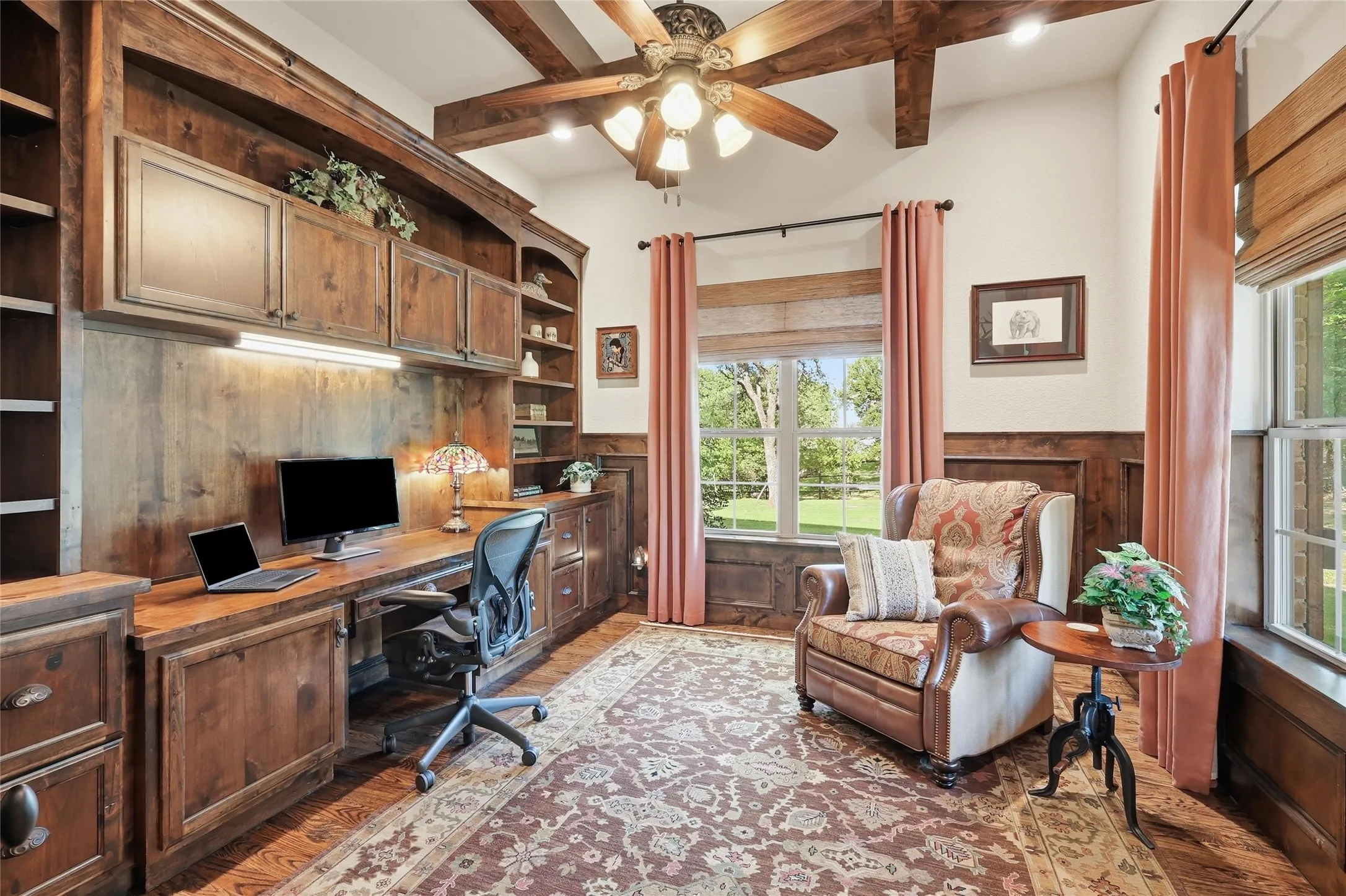 Study featuring extensive built-in custom cabinetry, wood-finish flooring, a ceiling fan, wood-paneled walls, and multiple windows