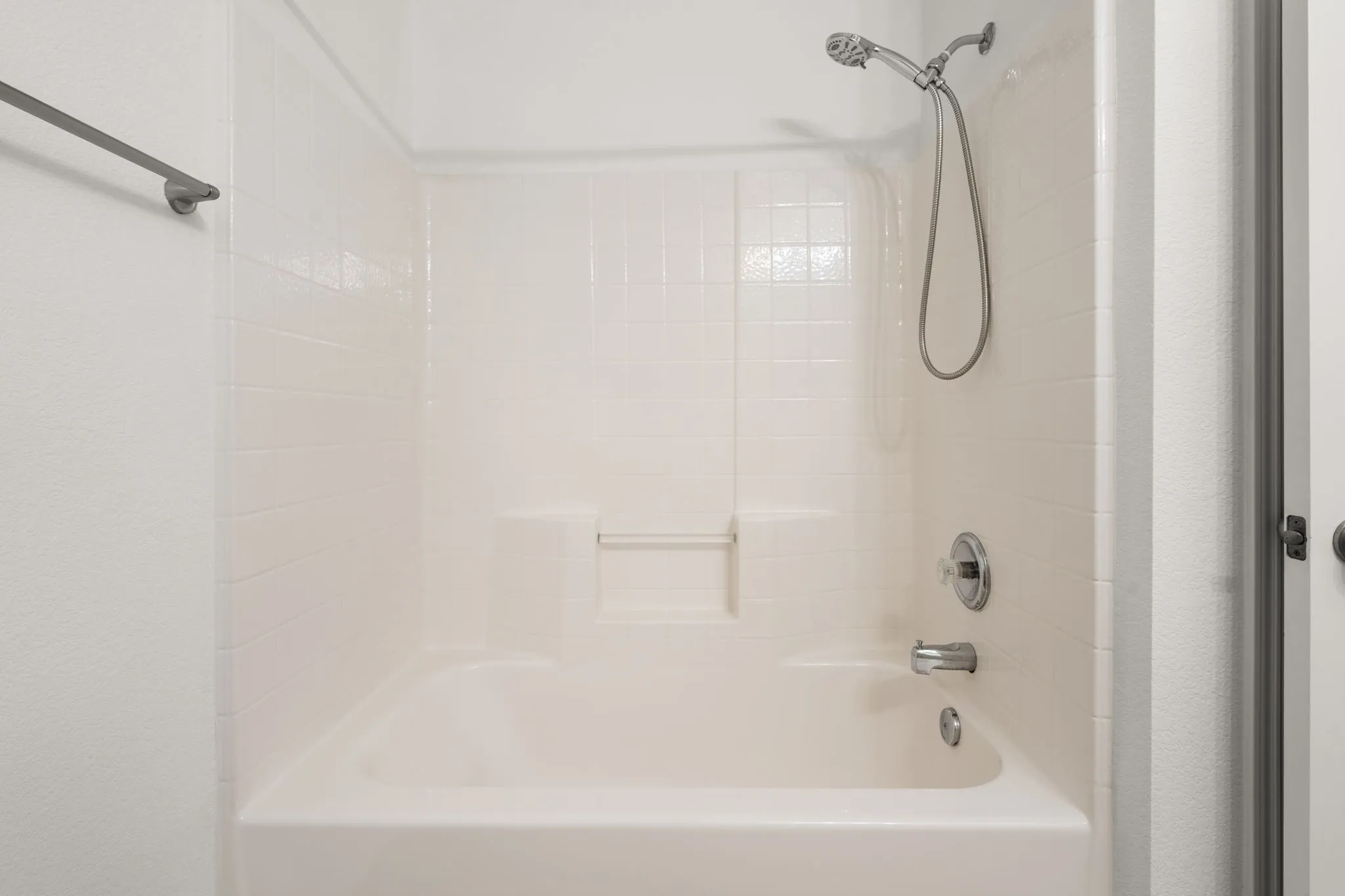 White tub and shower combination featuring a tiled surround, built-in shelving, and a handheld shower head with polished chrome fixtures