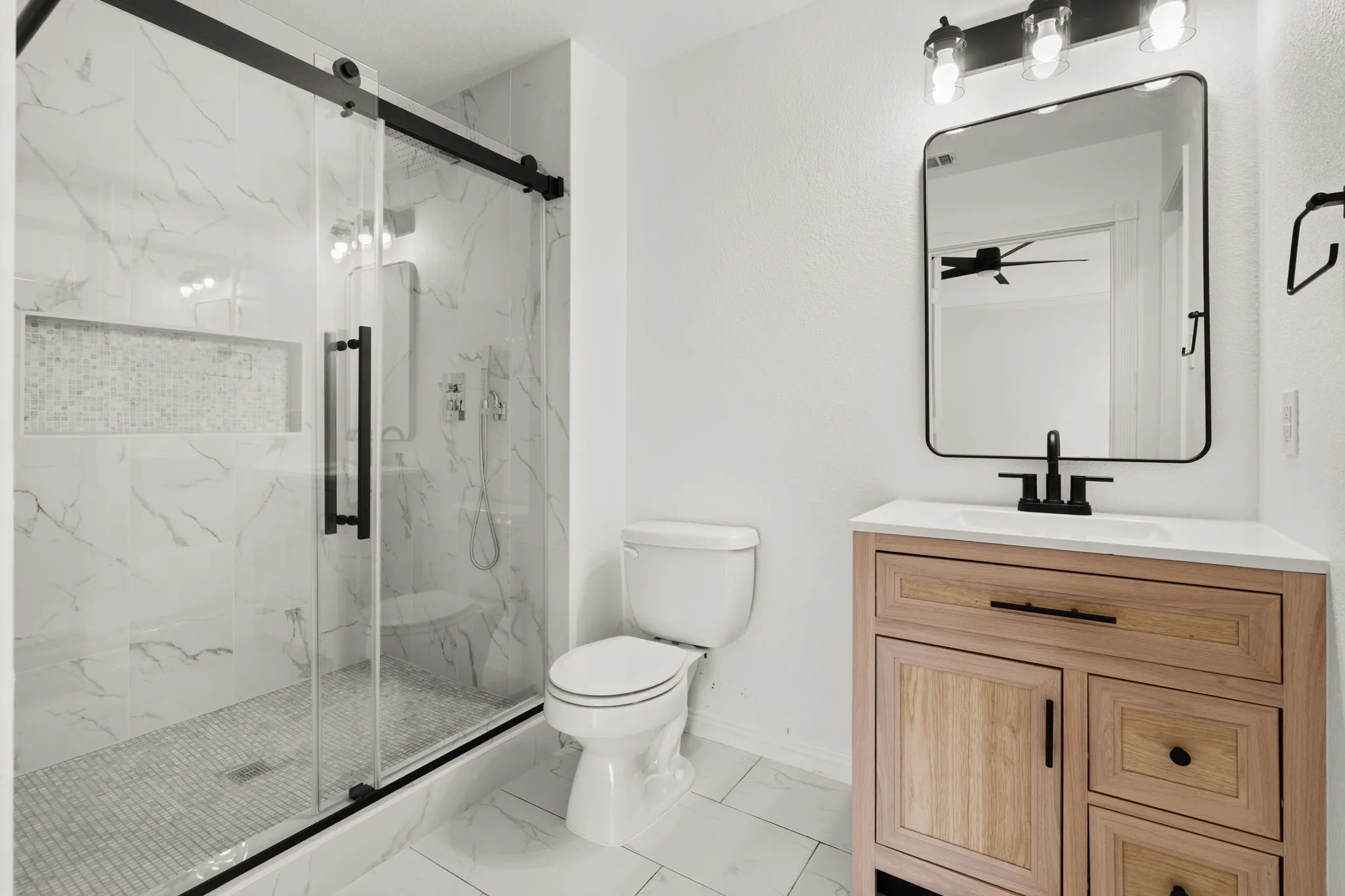 Modern bathroom featuring a glass-enclosed shower with black barn-door hardware, marble-look tile, and a mosaic-tiled shower niche