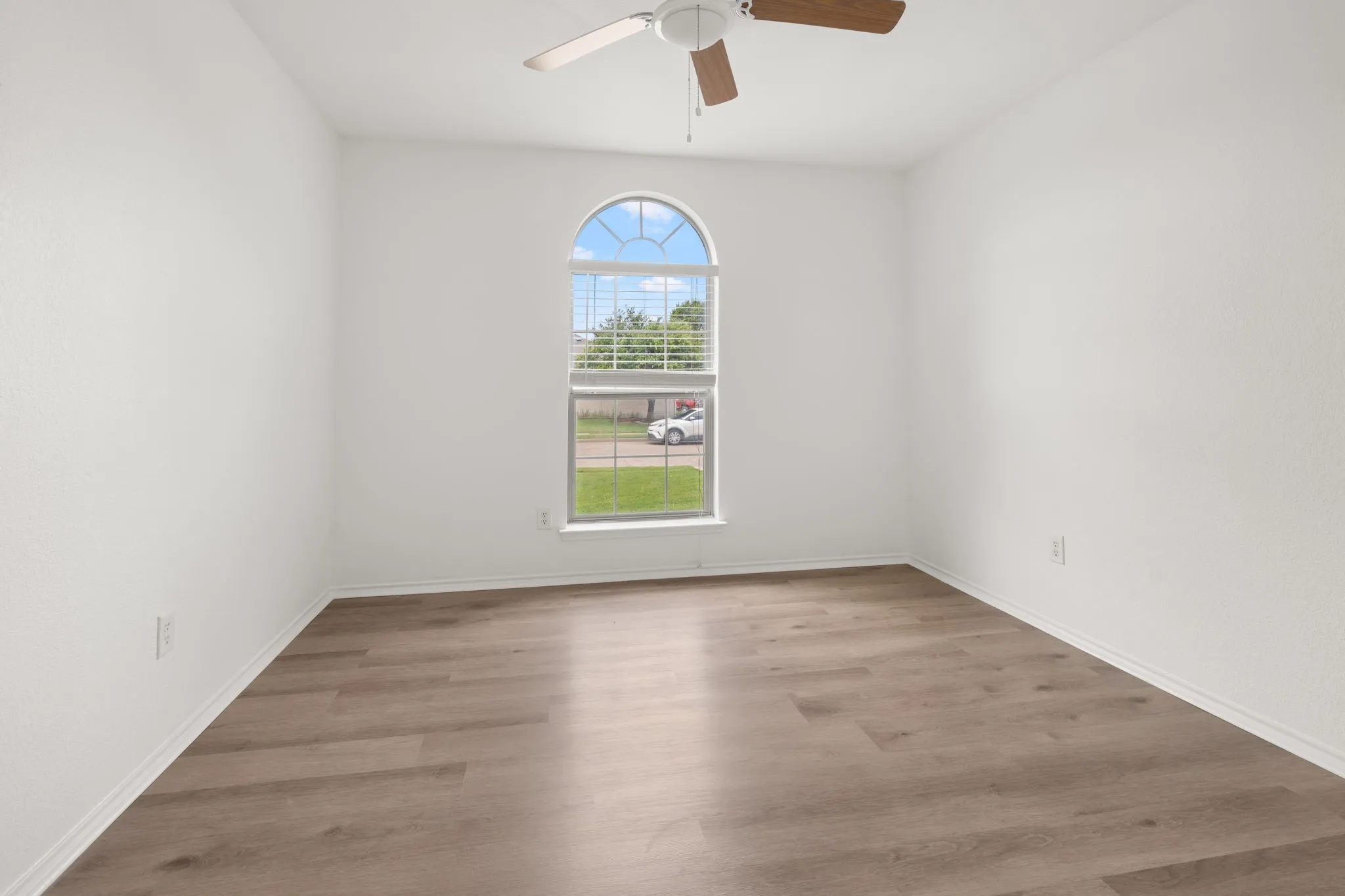 Room featuring a large arched window with horizontal blinds, wood-finish flooring, white walls, and a ceiling fan with wood-tone blades