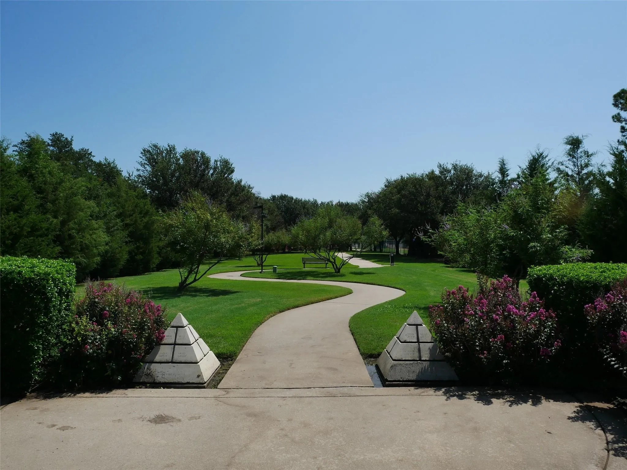 Expansive green space featuring a winding concrete pathway, mature trees, manicured hedges, flowering shrubs, and decorative pyramid-shaped stone accents