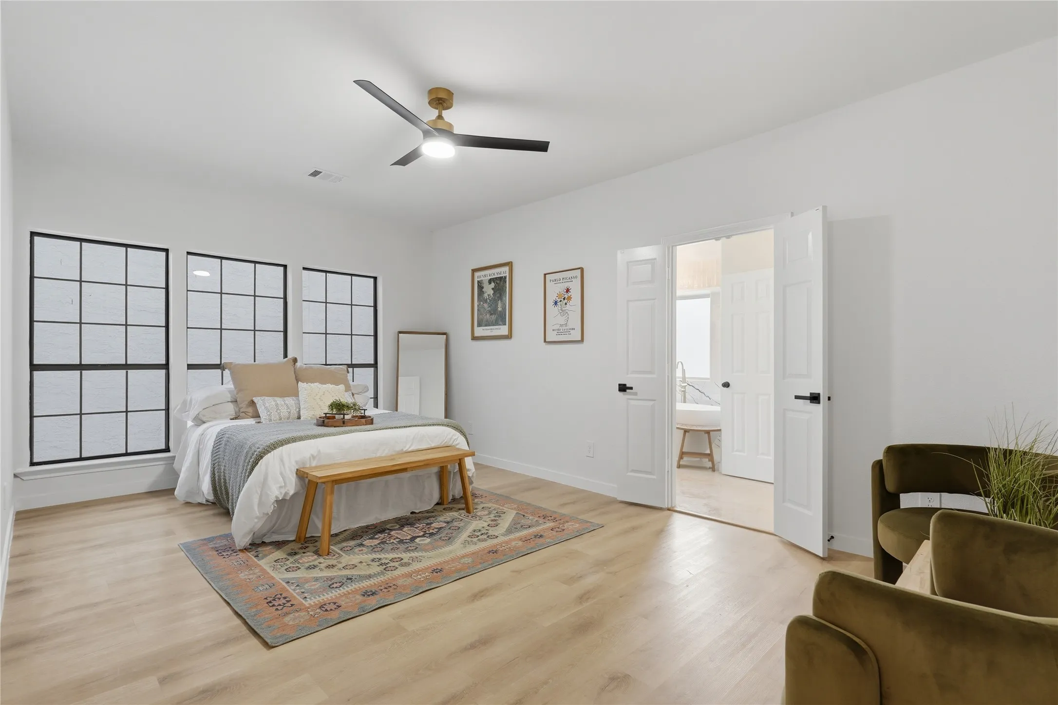 Spacious room featuring light wood-finish flooring, three grid-patterned windows with black frames, and a contemporary ceiling fan with a gold and black finish