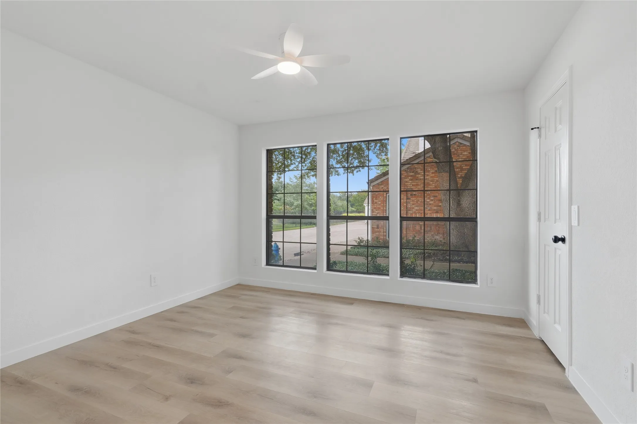 Bright room featuring three large multi-pane windows, wood-finish flooring, a contemporary ceiling fan with integrated lighting, white baseboards, and a paneled door with black hardware