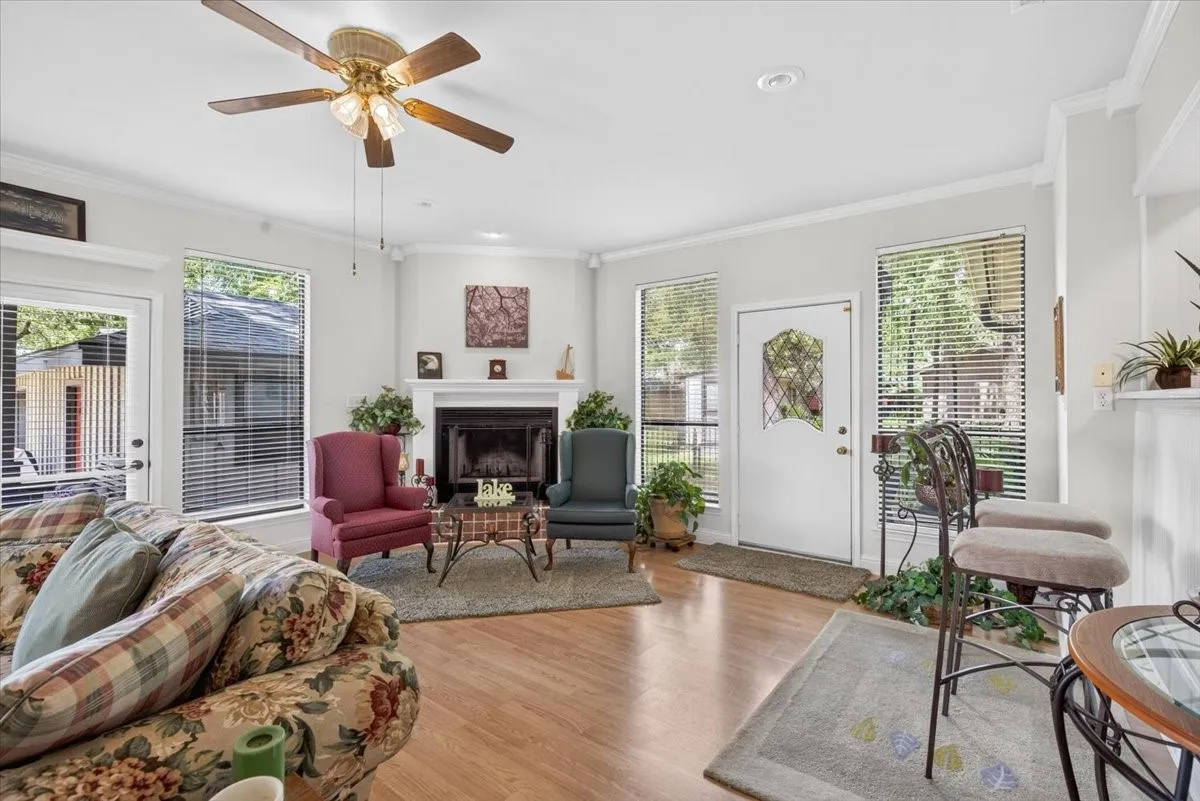 Living space featuring wood-finish flooring, a ceiling fan with integrated lighting, multiple windows with blinds, a white fireplace with a mantel, and a decorative front entry door