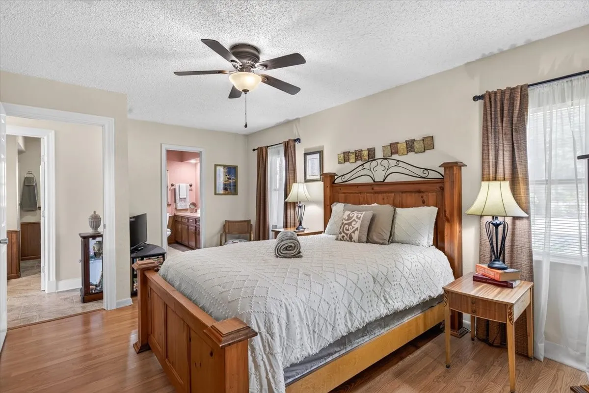 First floor Primary Bedroom featuring oak flooring, neutral colors, ceiling fan with integrated lighting, and multiple windows with blinds and ensuite bath.