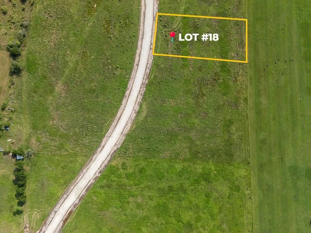 Expansive green lot #18, bordered by a gravel road, featuring open land and a level topography