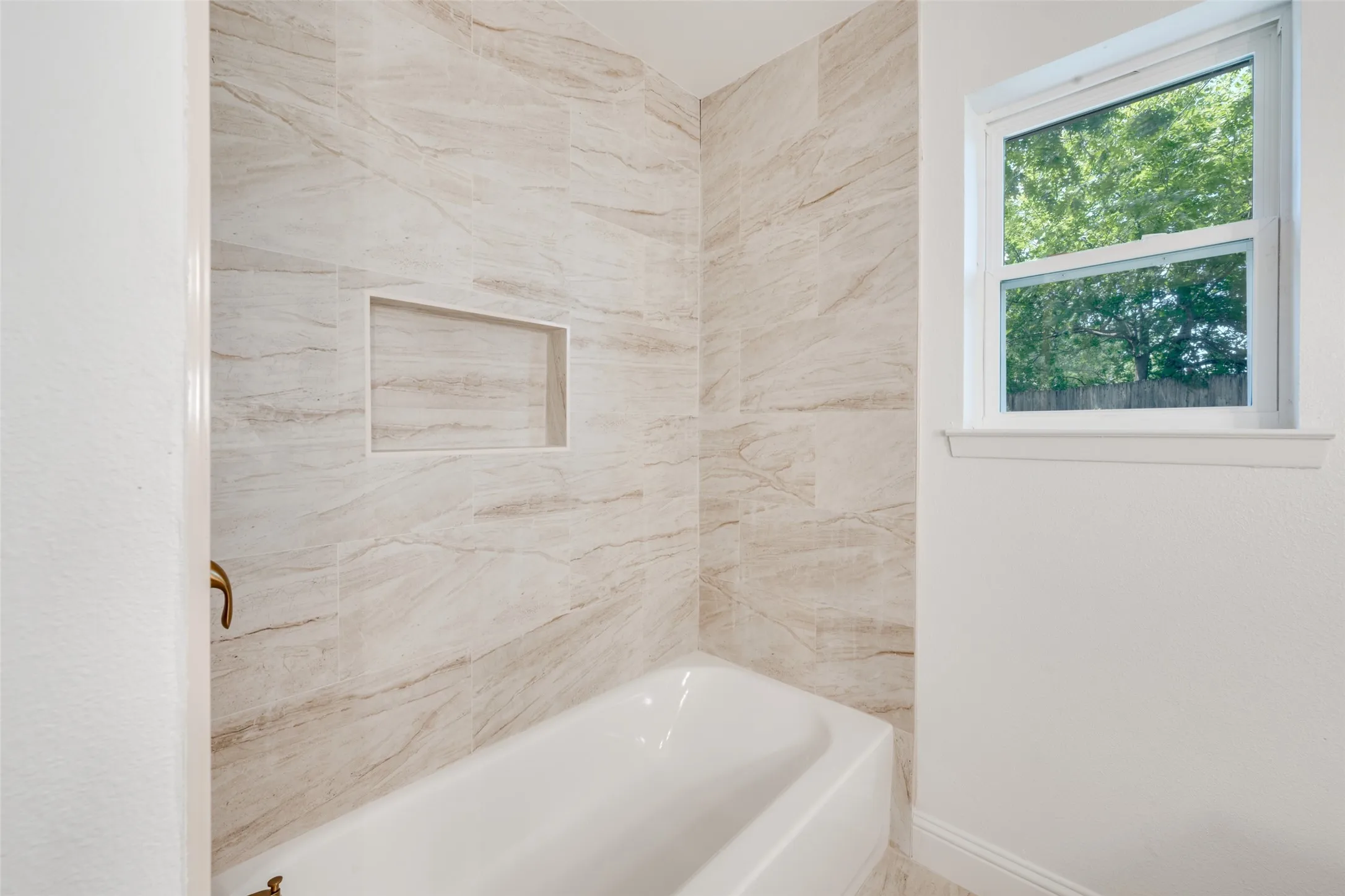 Bathtub and shower surround featuring large-format tile with natural stone-look pattern, an integrated shower niche, and a white framed window