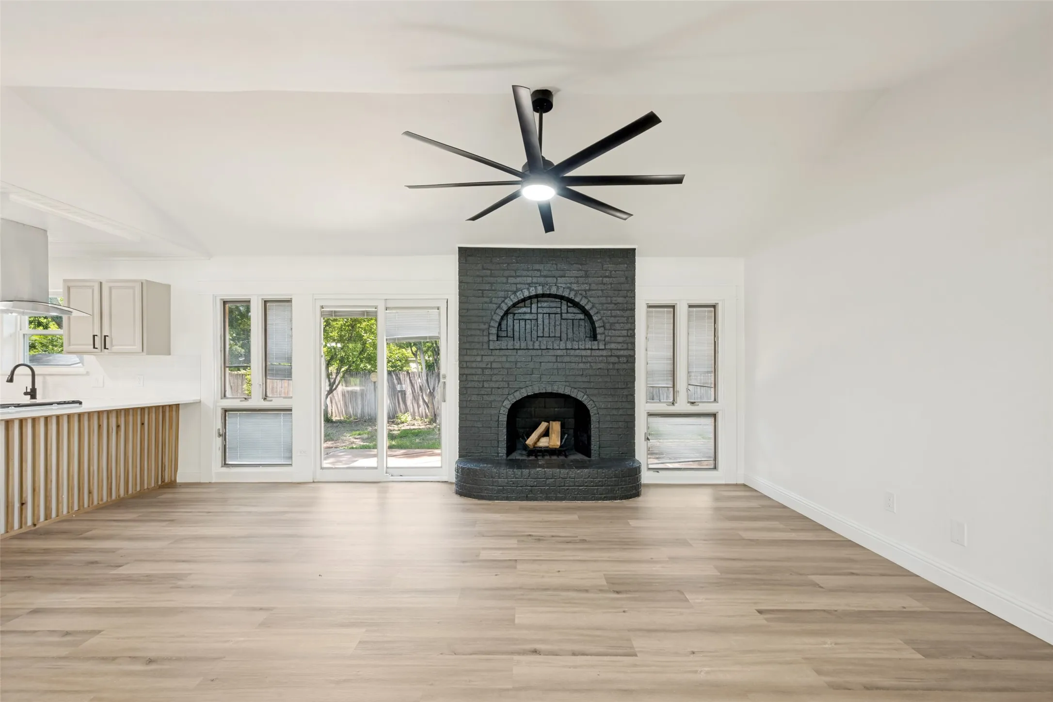 Vaulted ceiling with a contemporary black ceiling fan, dark brick fireplace with an arched detail, wood-finish flooring, white walls, and a sliding glass door opening to the outdoors