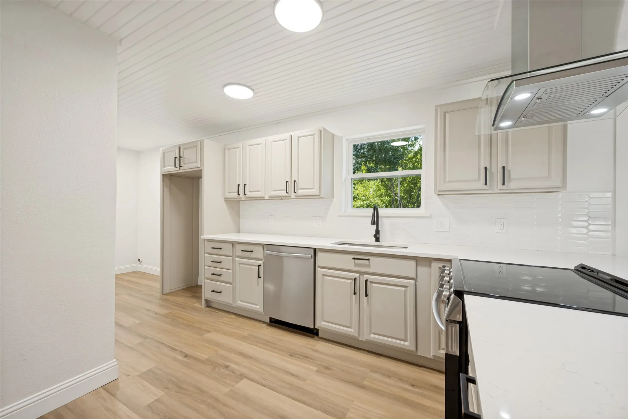 Modern kitchen featuring light cabinetry, wood-finish flooring, white subway tile backsplash, stainless steel appliances, and a range hood