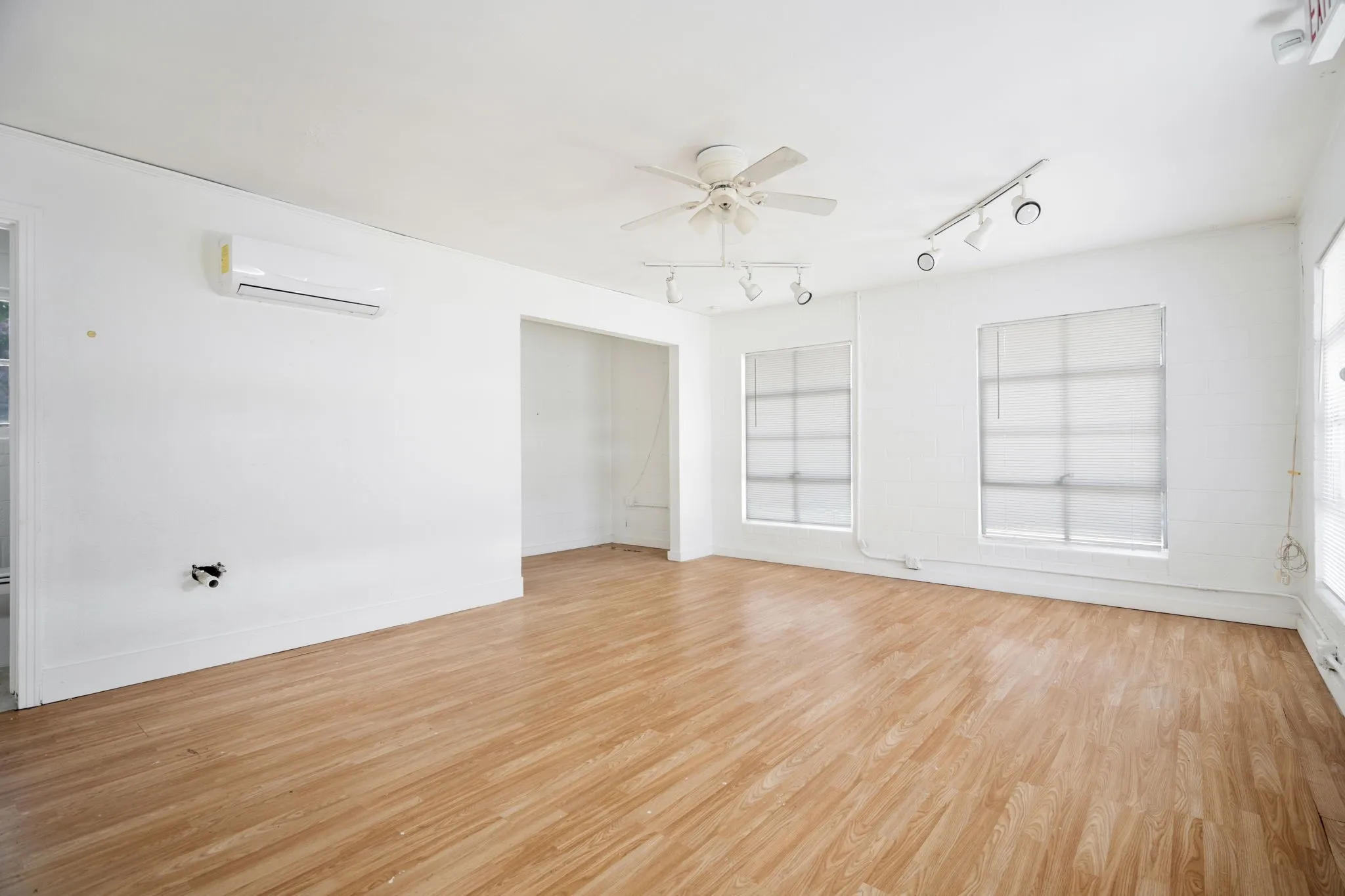 Spacious room featuring wood-finish flooring, white walls, multiple windows with blinds, track lighting, and a ceiling fan