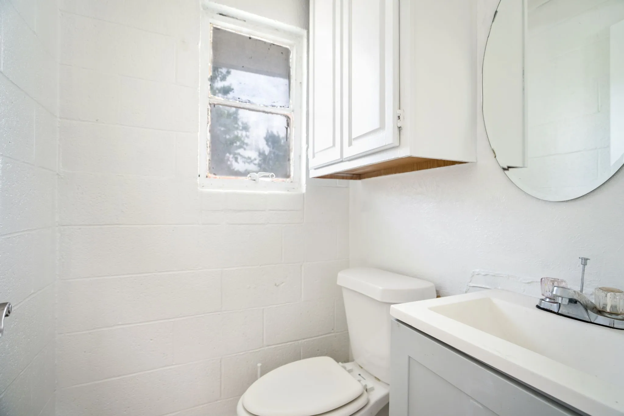 Bathroom featuring a window, white painted block walls, a toilet, a vanity with a white sink, and a circular mirror