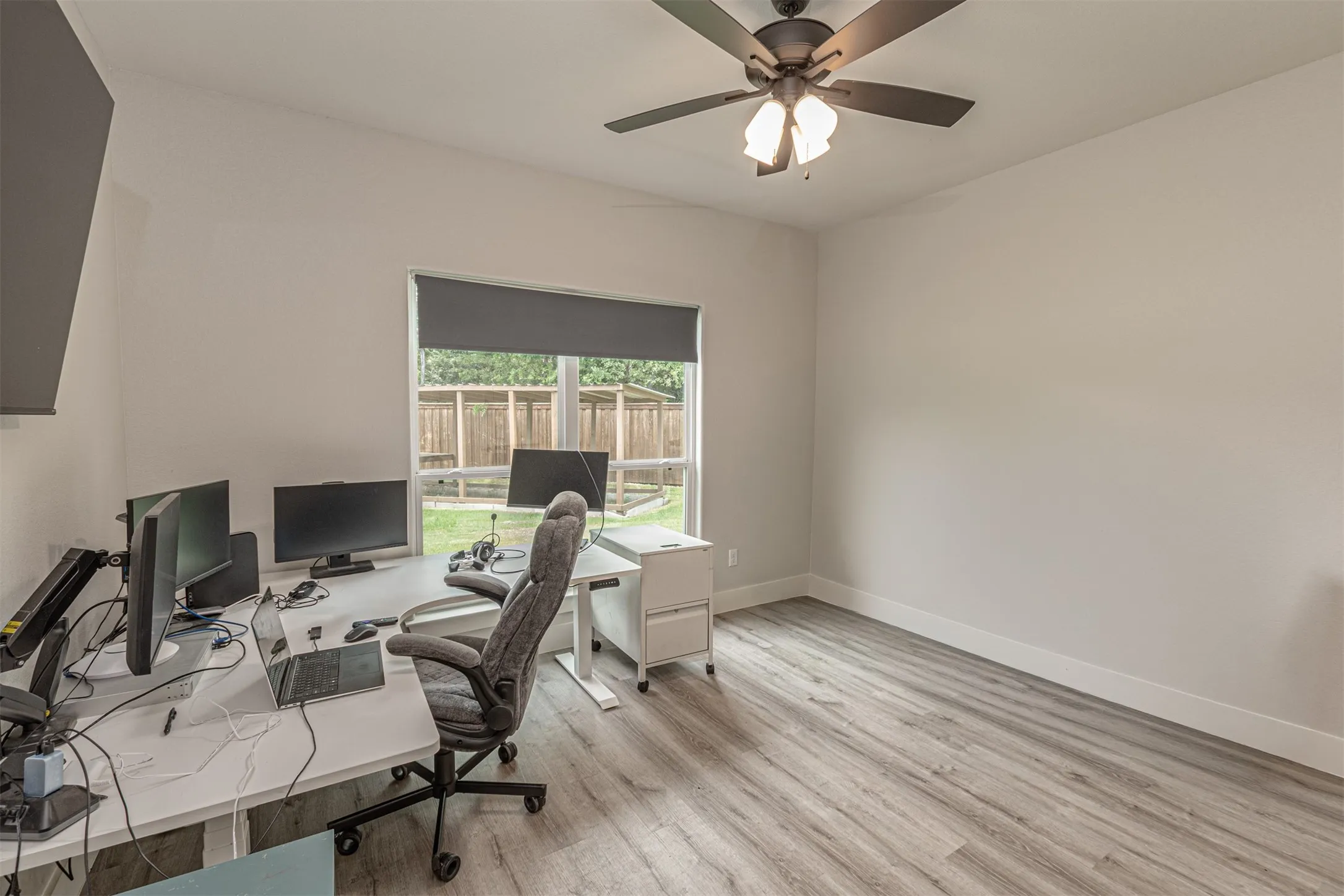 Neutral-toned room featuring wood-finish flooring, a contemporary ceiling fan with integrated lighting, and a large window with a roller shade overlooking a fenced exterior