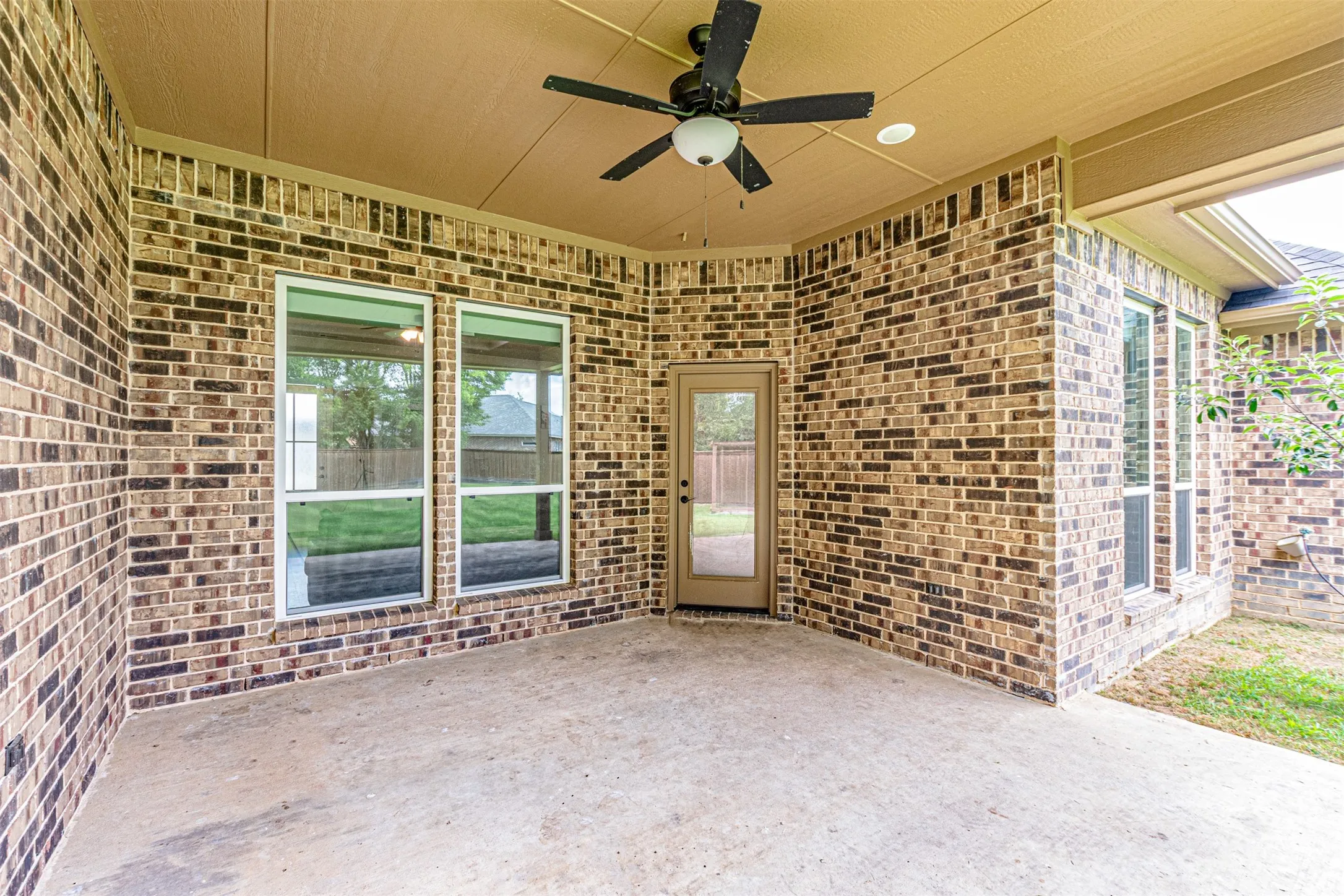 Covered concrete patio featuring a ceiling fan with integrated lighting, brick exterior walls, and multiple windows