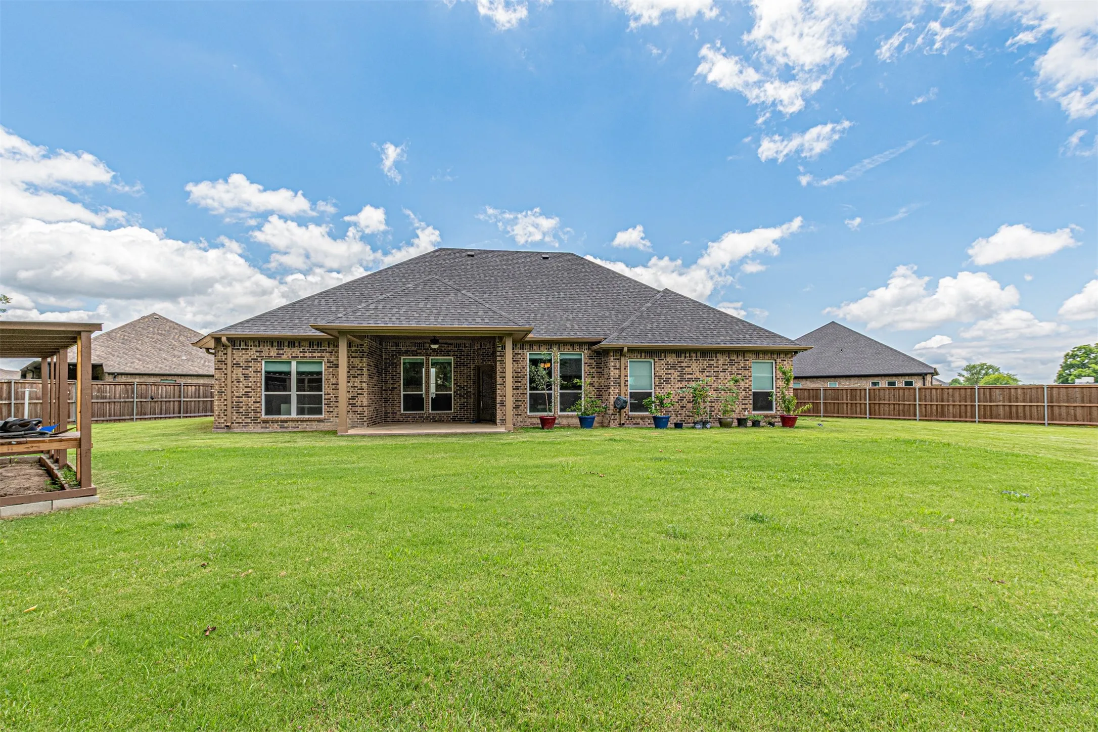 Expansive rear lawn featuring a brick exterior, covered patio, dark shingle roof, and a wood privacy fence
