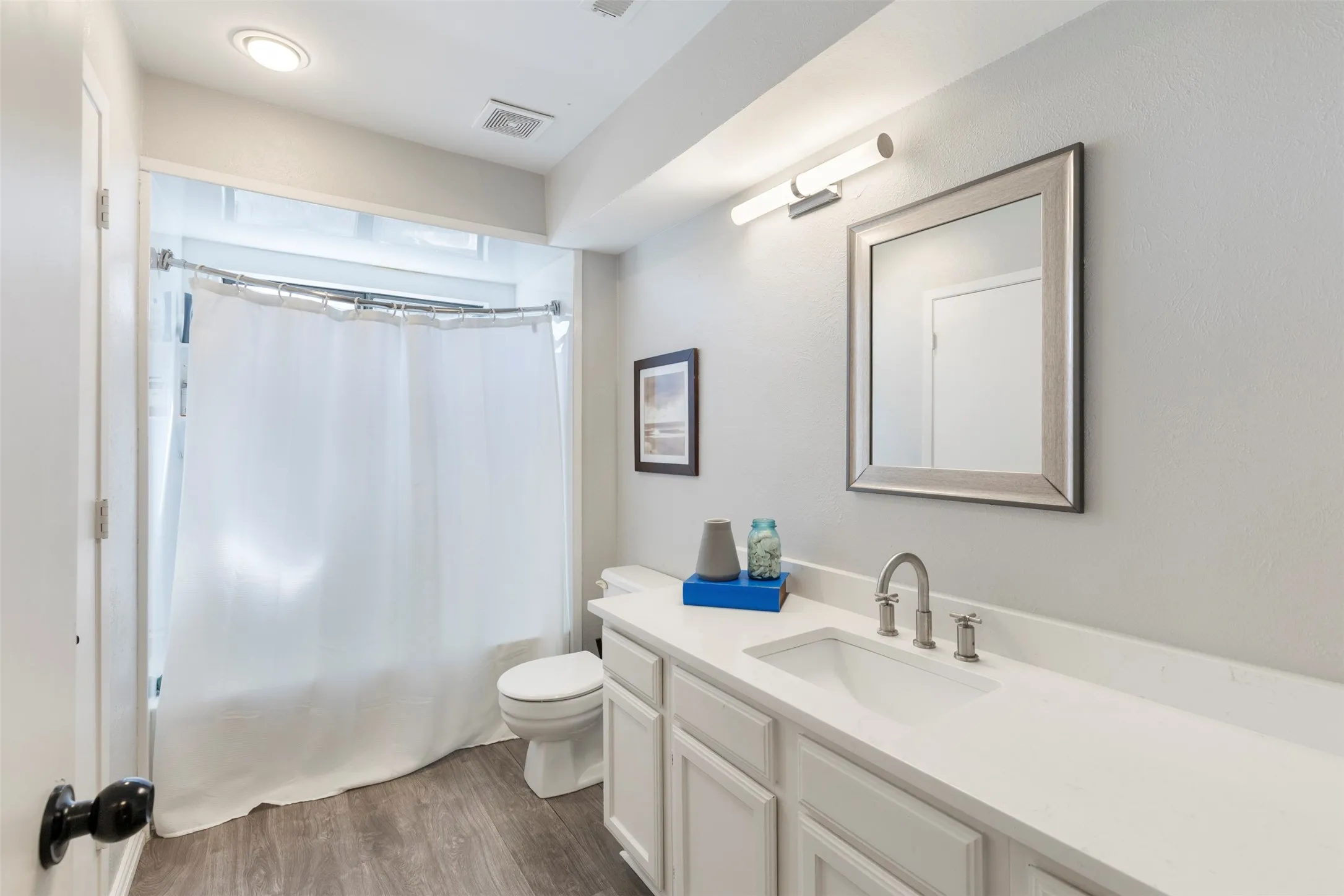 Full bathroom featuring a white vanity with an integrated sink, silver-framed mirror, and wood-finish flooring