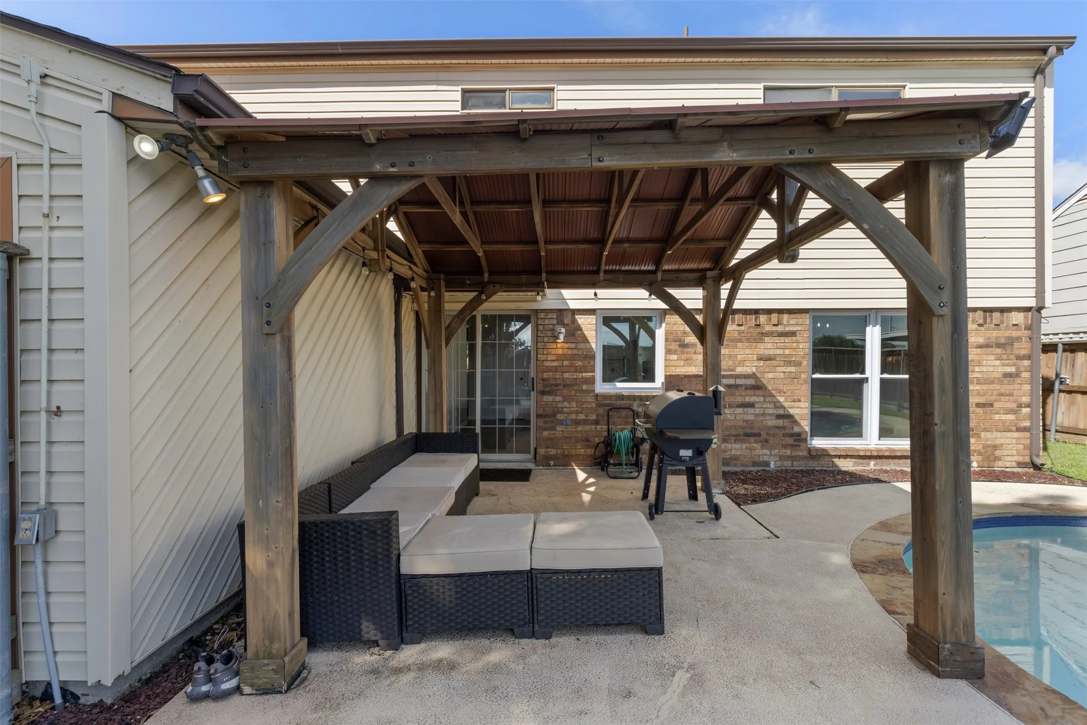 Expansive outdoor patio featuring a wooden pergola with a corrugated roof, concrete hardscaping, and direct access to a swimming pool