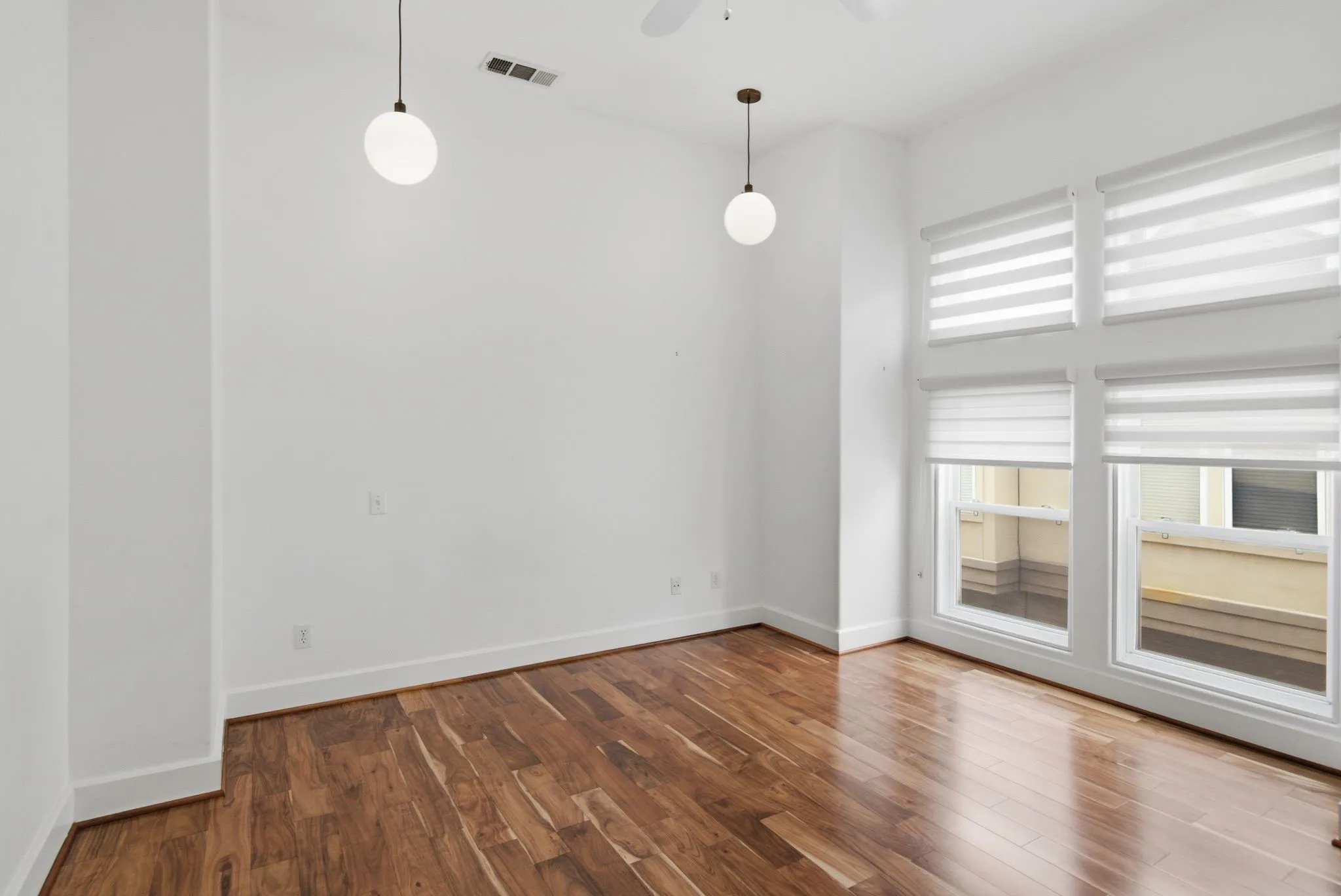 Spacious room featuring rich wood-finish flooring, high ceilings, multiple tall windows with integrated blinds, and contemporary spherical pendant lighting