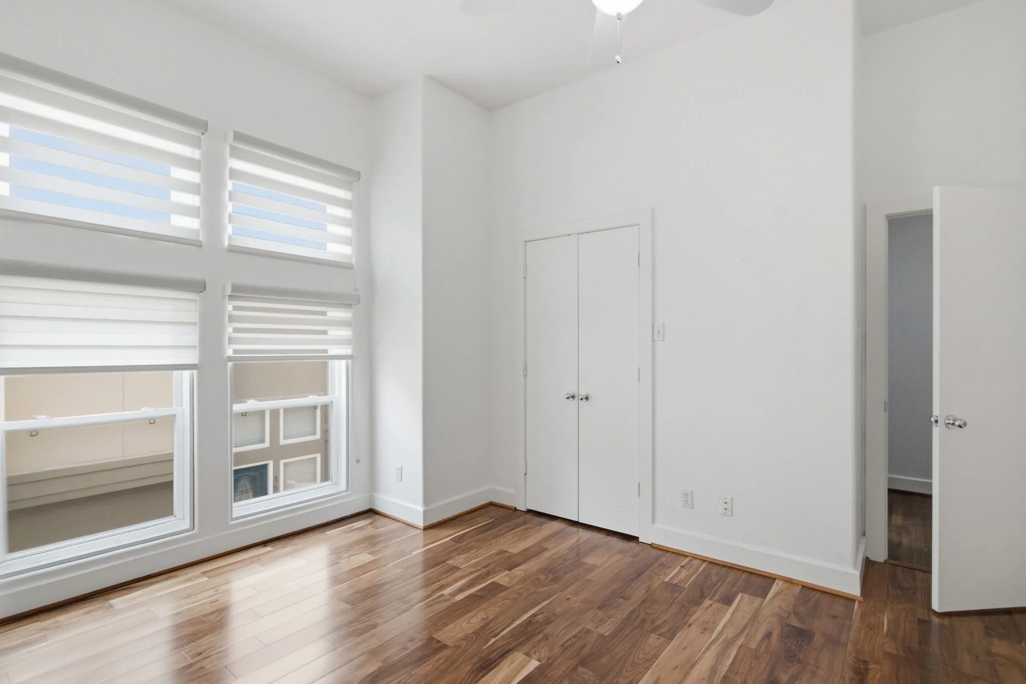 Hardwood-finish flooring, high ceilings, multiple windows with blinds, built-in closet with double doors, and a ceiling fan with light fixture