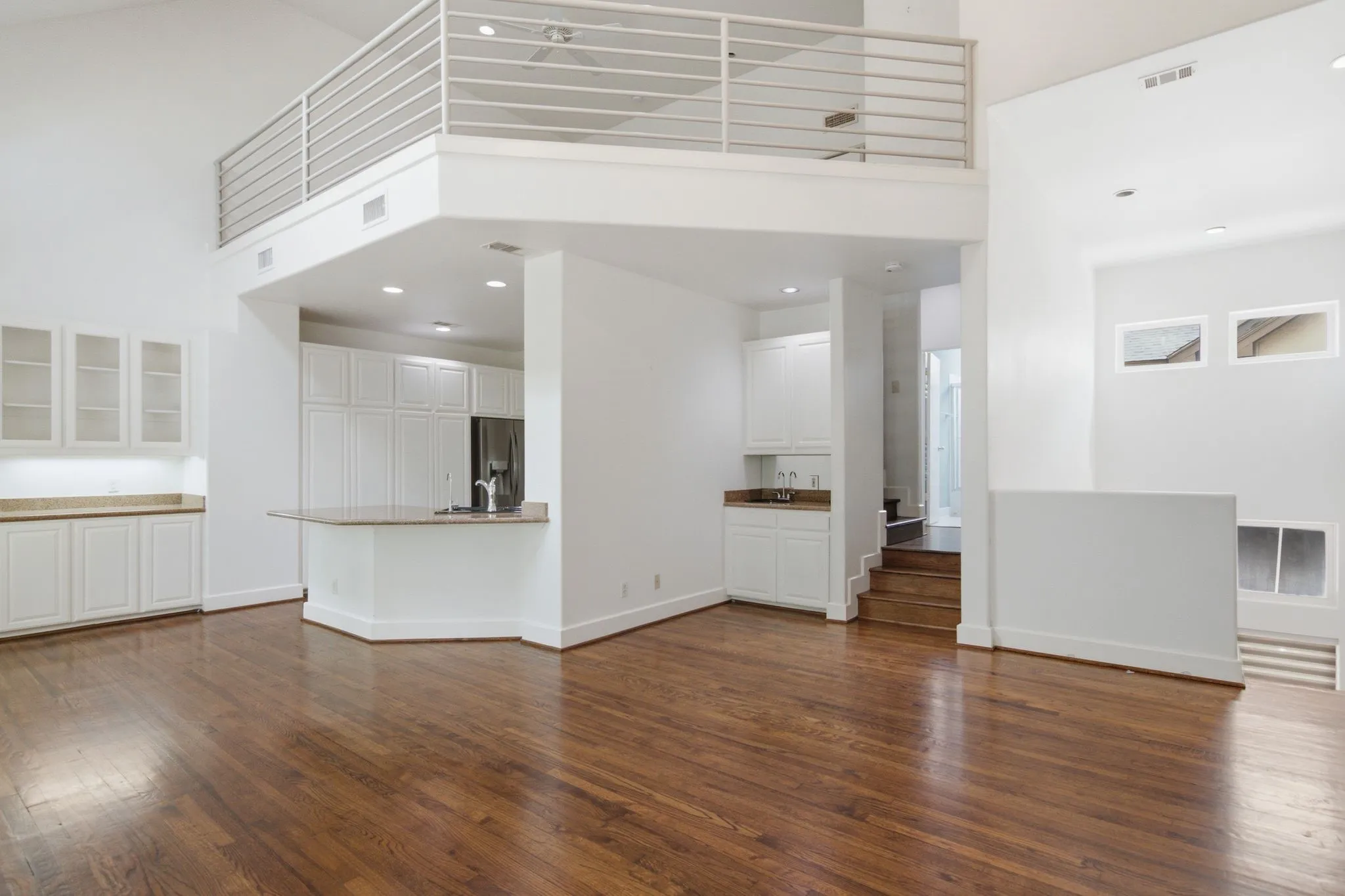 Spacious living area featuring polished wood flooring, high ceilings, a loft with metal railing, built-in cabinetry with glass inserts, and a kitchen island with stone countertops