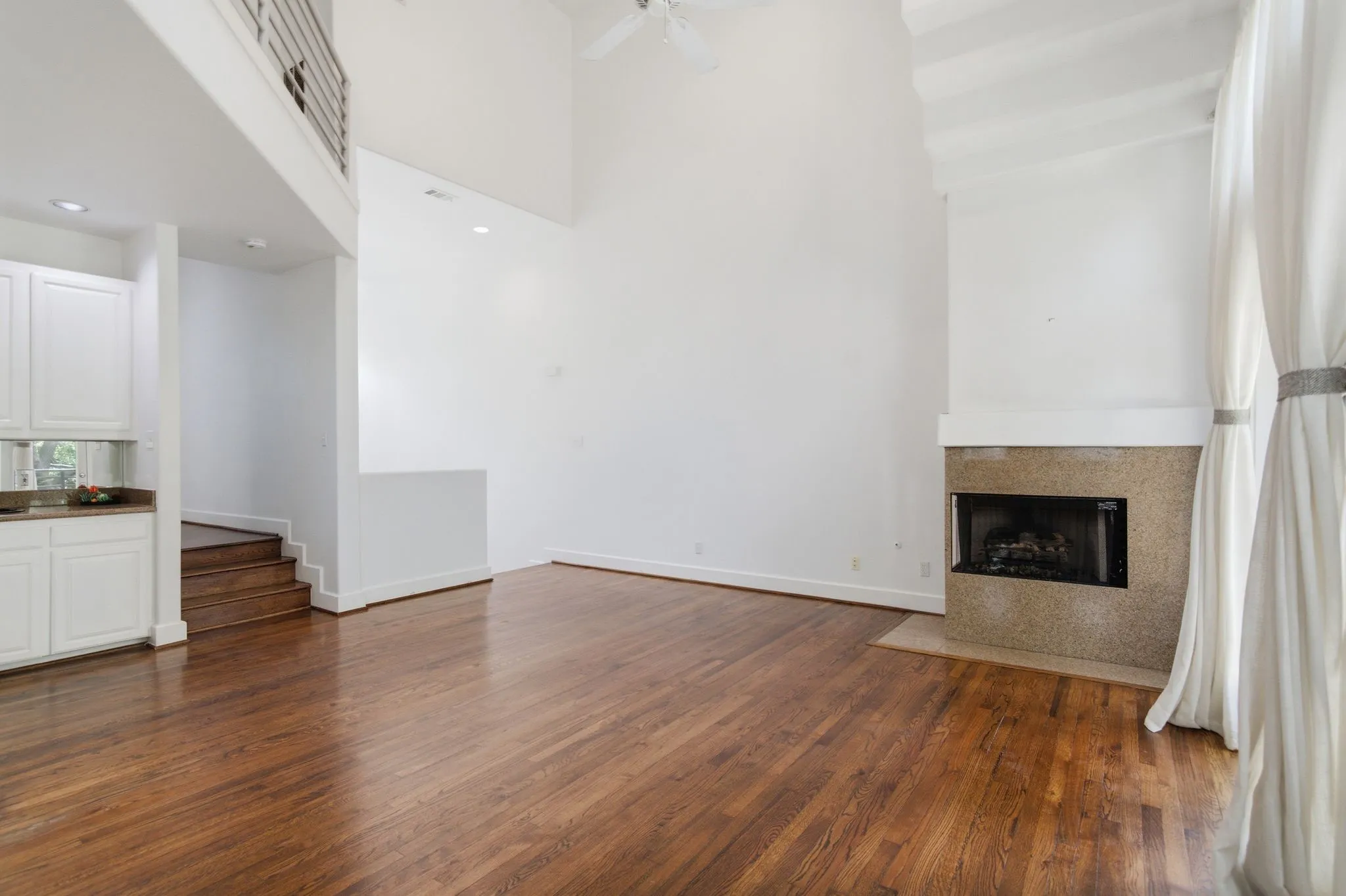 Spacious living area featuring high ceilings, wood-finish flooring, a built-in fireplace with a stone surround, and white floor-to-ceiling drapery