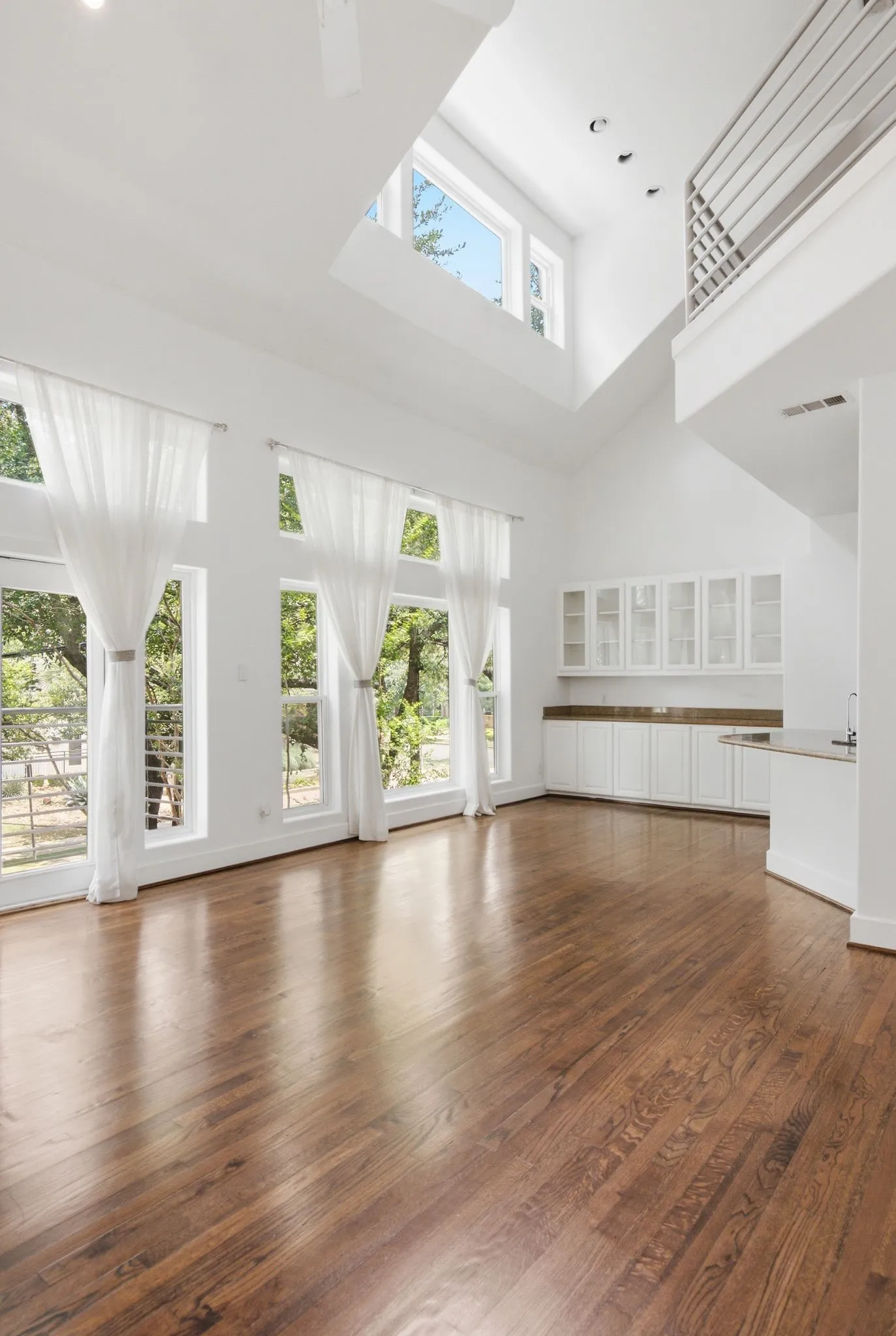 Vaulted ceiling great room featuring expansive windows, wood-finish flooring, a built-in wet bar, and an upper-level balcony with metal railing