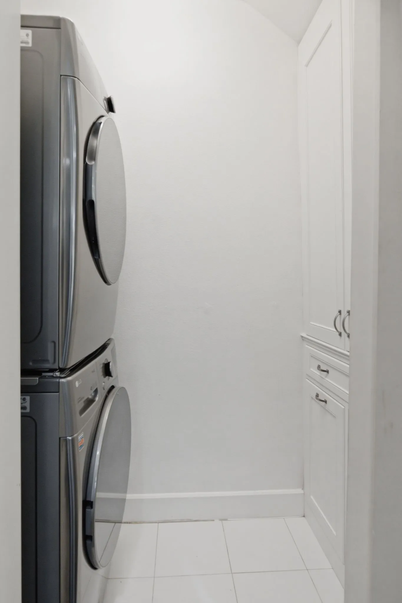 Dedicated laundry area featuring a stacked washer and dryer, white tile flooring, white baseboards, and built-in white cabinetry with chrome hardware